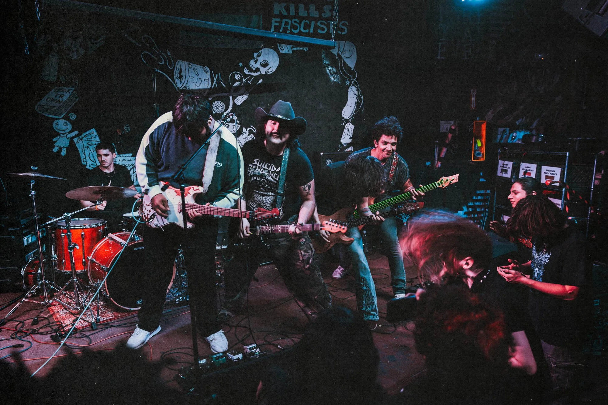 A live band performance on stage with five band members playing guitars and drums, in a dimly lit venue with graffiti-style artwork on the back wall.