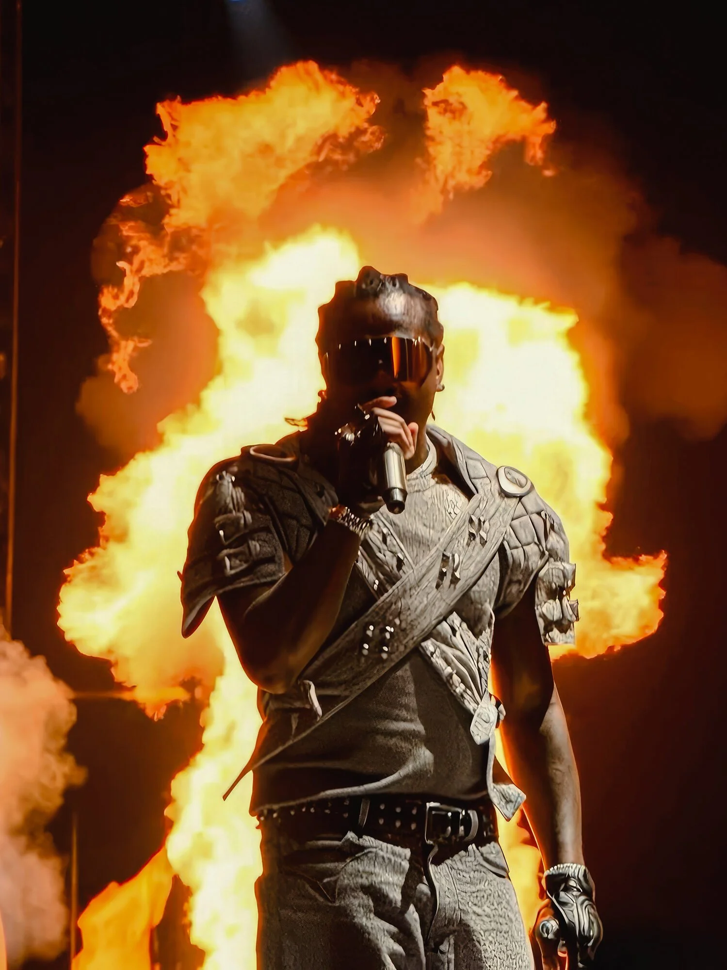 A person wearing sunglasses and a tactical outfit standing in front of a large fire explosion at night, holding a microphone.