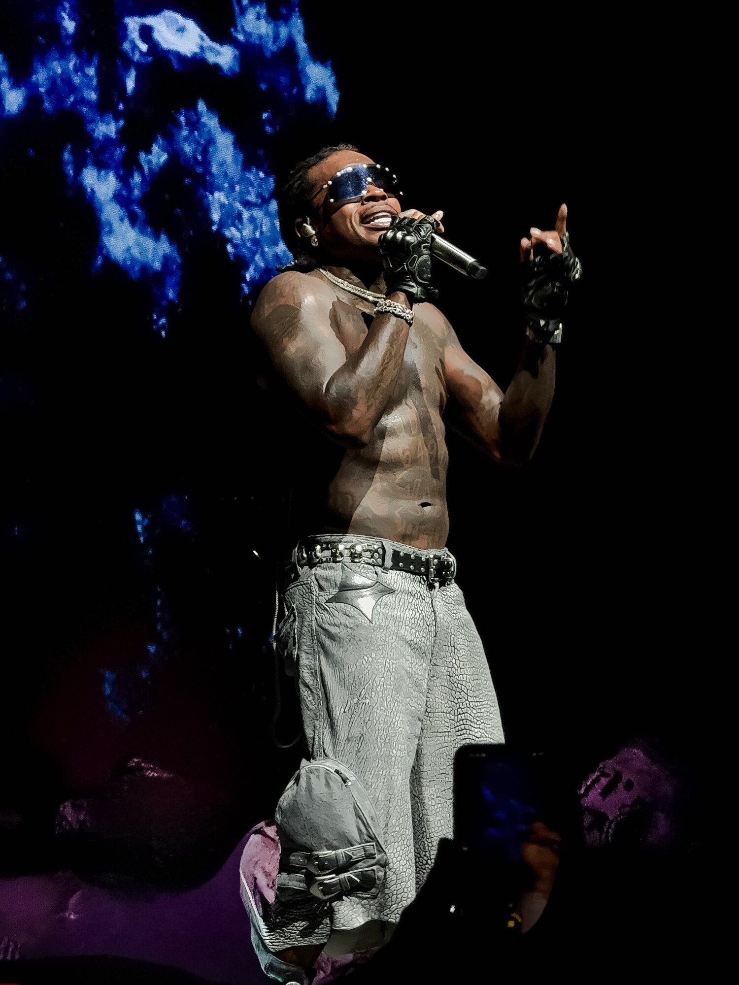 A shirtless male performer with tattoos, wearing sunglasses, gloves, and baggy jeans, singing into a microphone on stage with a dark background. Gunna concert.