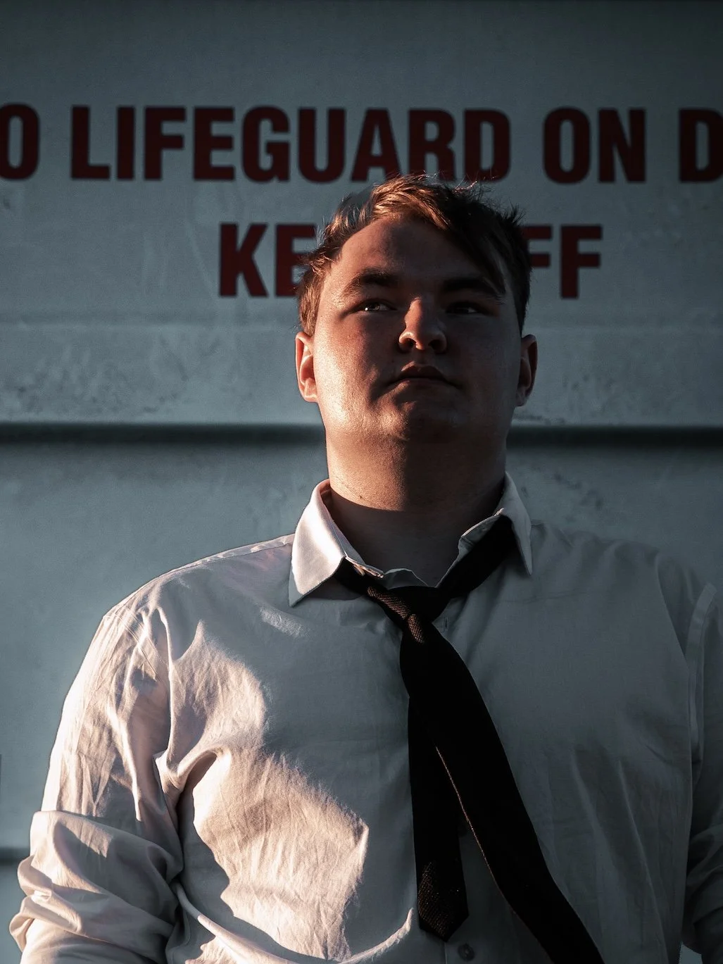 Young man in a white shirt and black tie standing in front of a wall with a warning sign that reads 'No Lifeguard on Duty'.