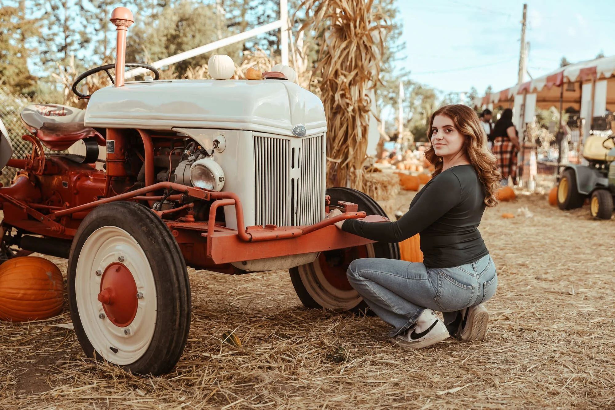 A young woman kneeling next to a vintage white and red tractor at a pumpkin patch or fall festival, with pumpkins and hay in the background.