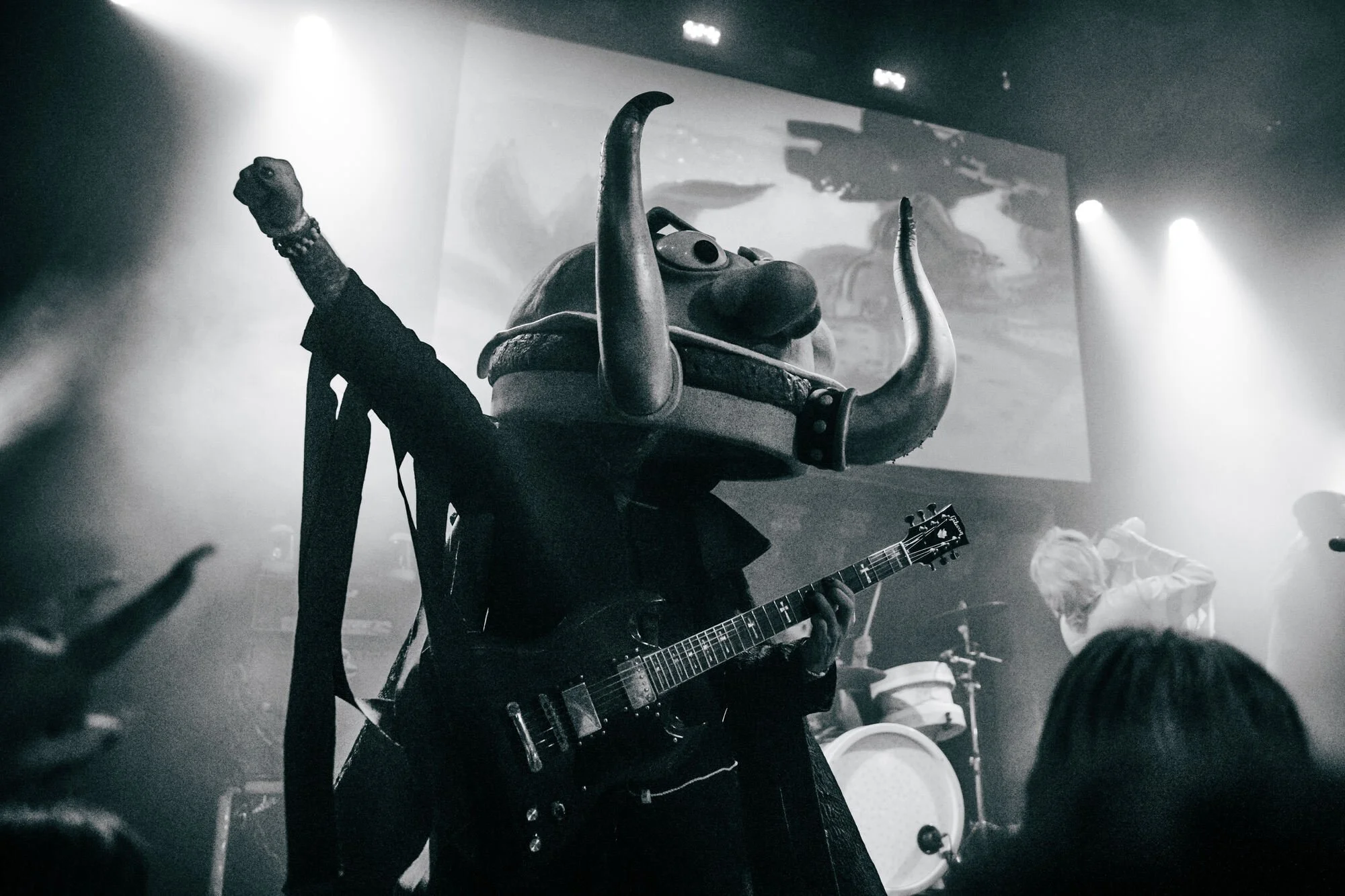 Person wearing a large, animal-shaped mask with horns, playing an electric guitar on stage at a concert.