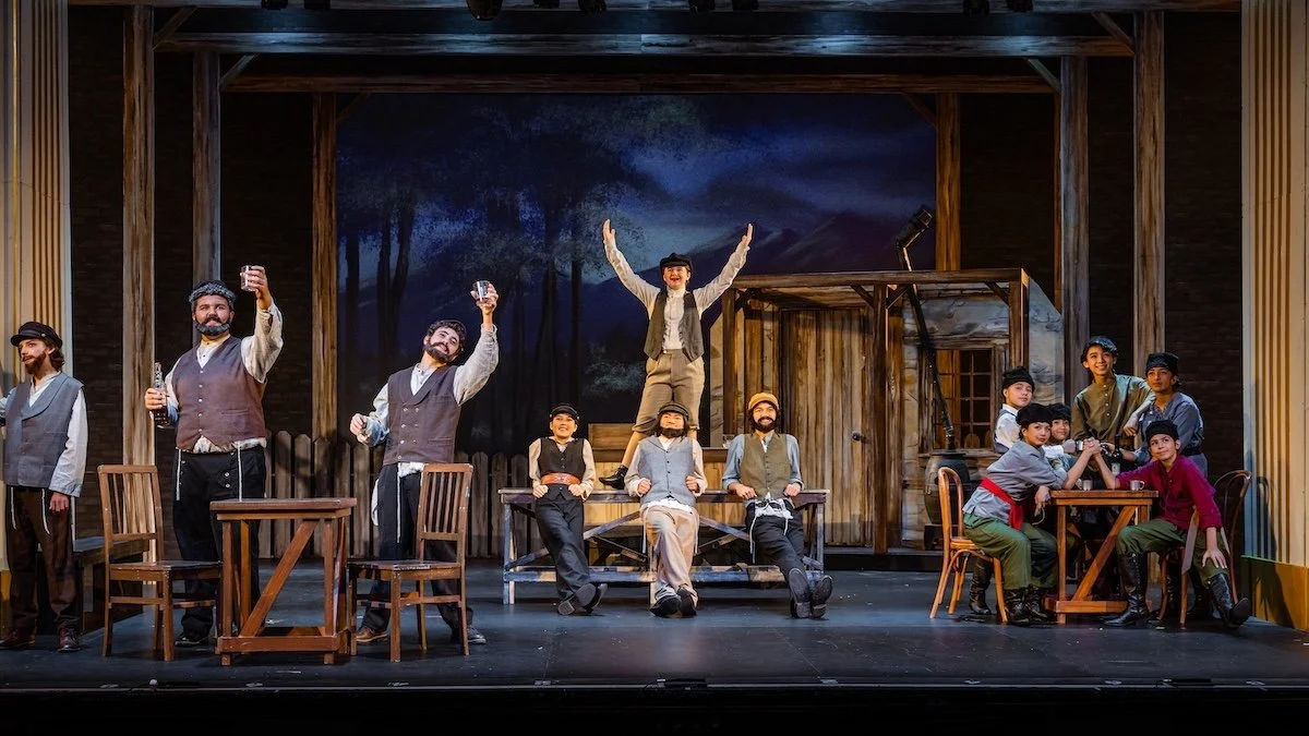 The scene shows a theatrical stage with actors in period costumes, some standing, sitting, or raising glasses, and one actor balancing on a platform with arms outstretched. The set resembles an outdoor rustic setting with a wooden building and trees 