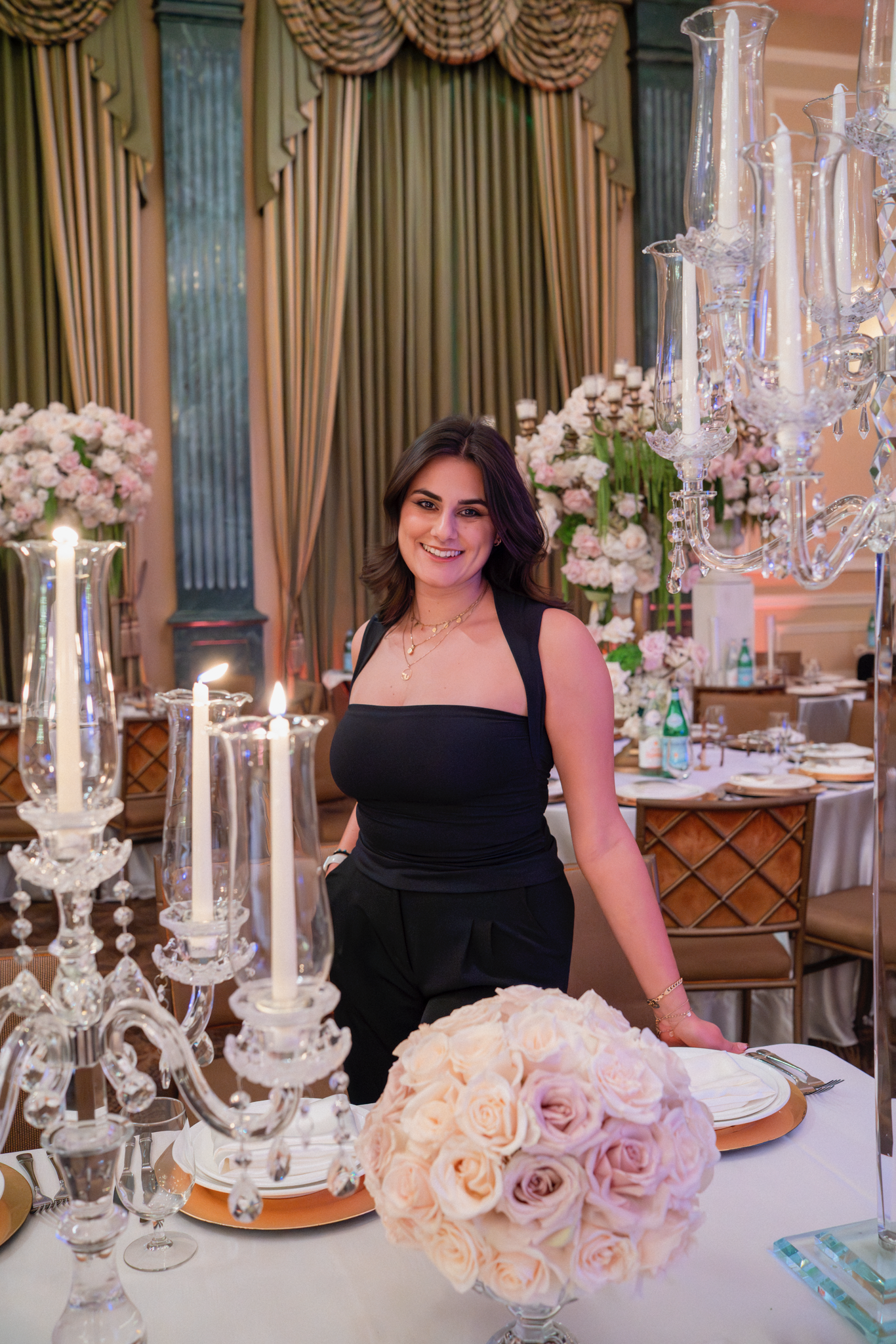 A woman in a black dress smiling at a decorated banquet table with floral arrangements and crystal candelabras, in a formal event setting.