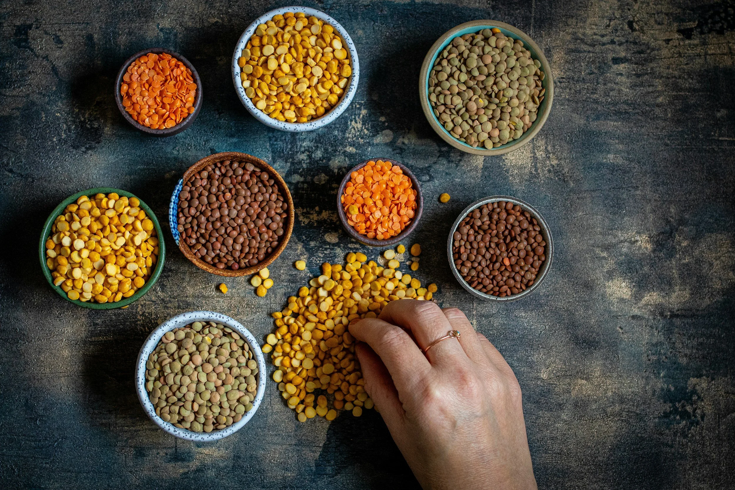 A hand picking up yellow split peas from a small dish, with multiple small bowls of lentils and split peas in various colors on a dark textured surface.