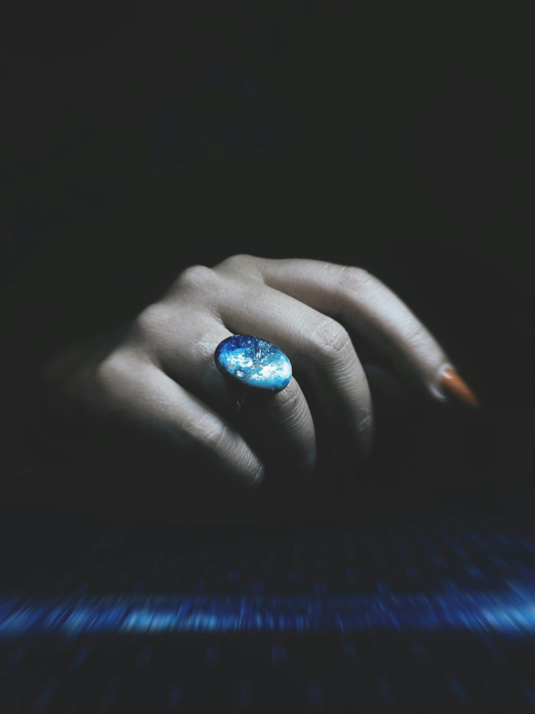 A close-up of a person's hand with a large oval-shaped blue galaxy-themed ring on their finger, against a dark background.