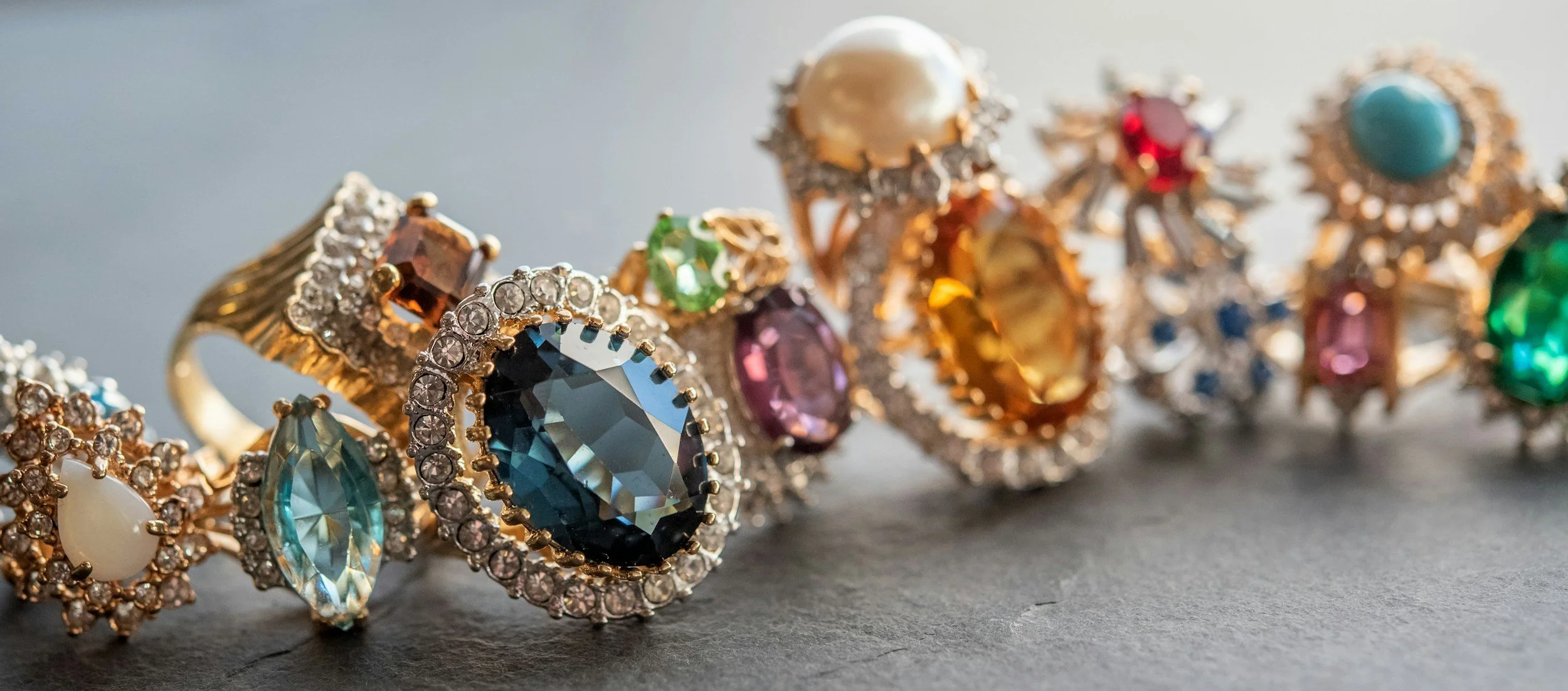 Close-up of various colorful gemstone rings with intricate designs on a gray surface.