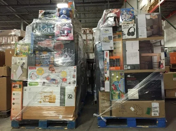 Stacks of boxed household products and toys wrapped in plastic on pallets in a warehouse.