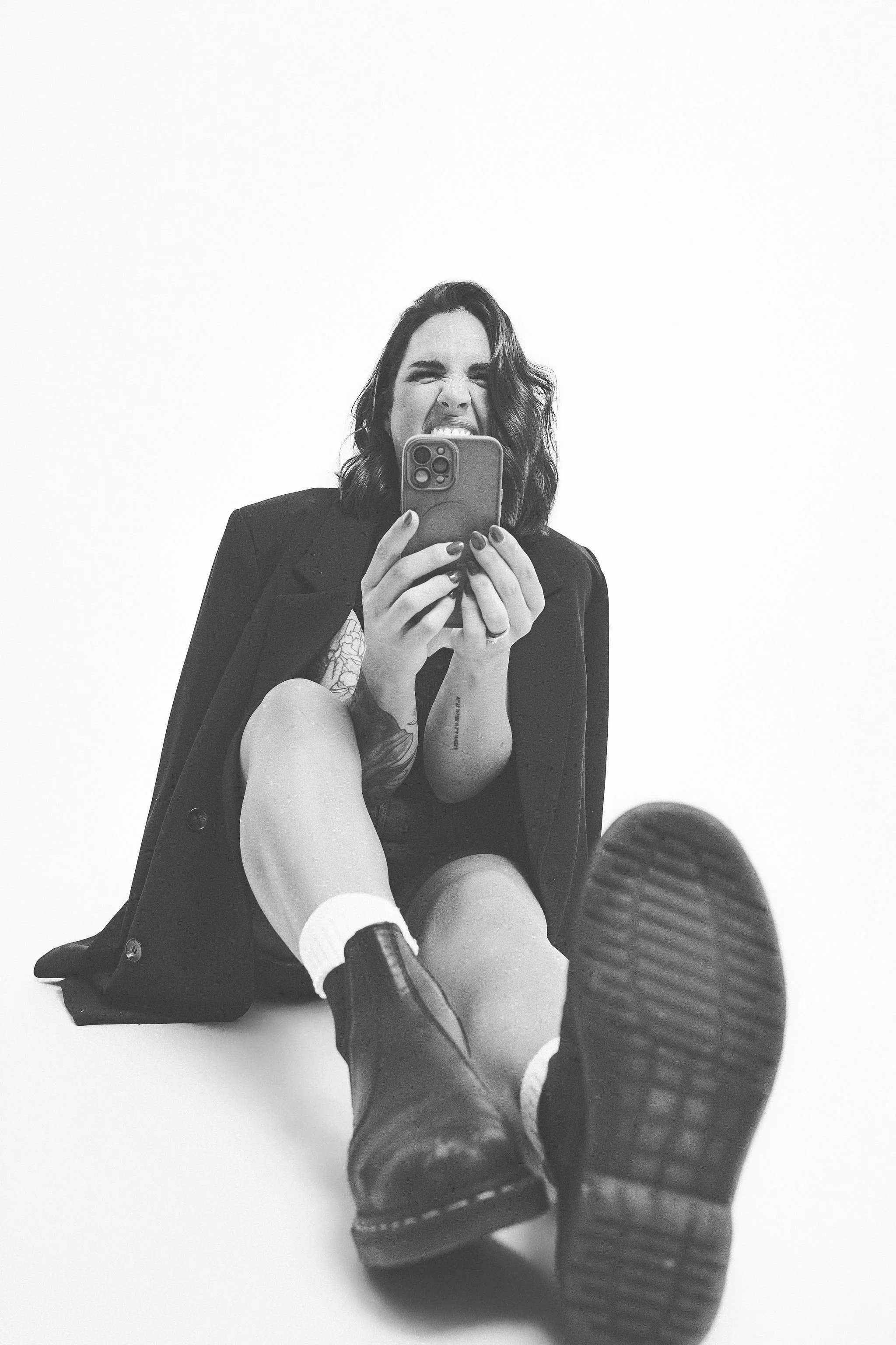 Black and white photo of a woman sitting on the floor, taking a selfie with her phone, smiling and making a playful expression, dressed in a black jacket, with tattoos visible on her arms and leg, wearing boots and socks.