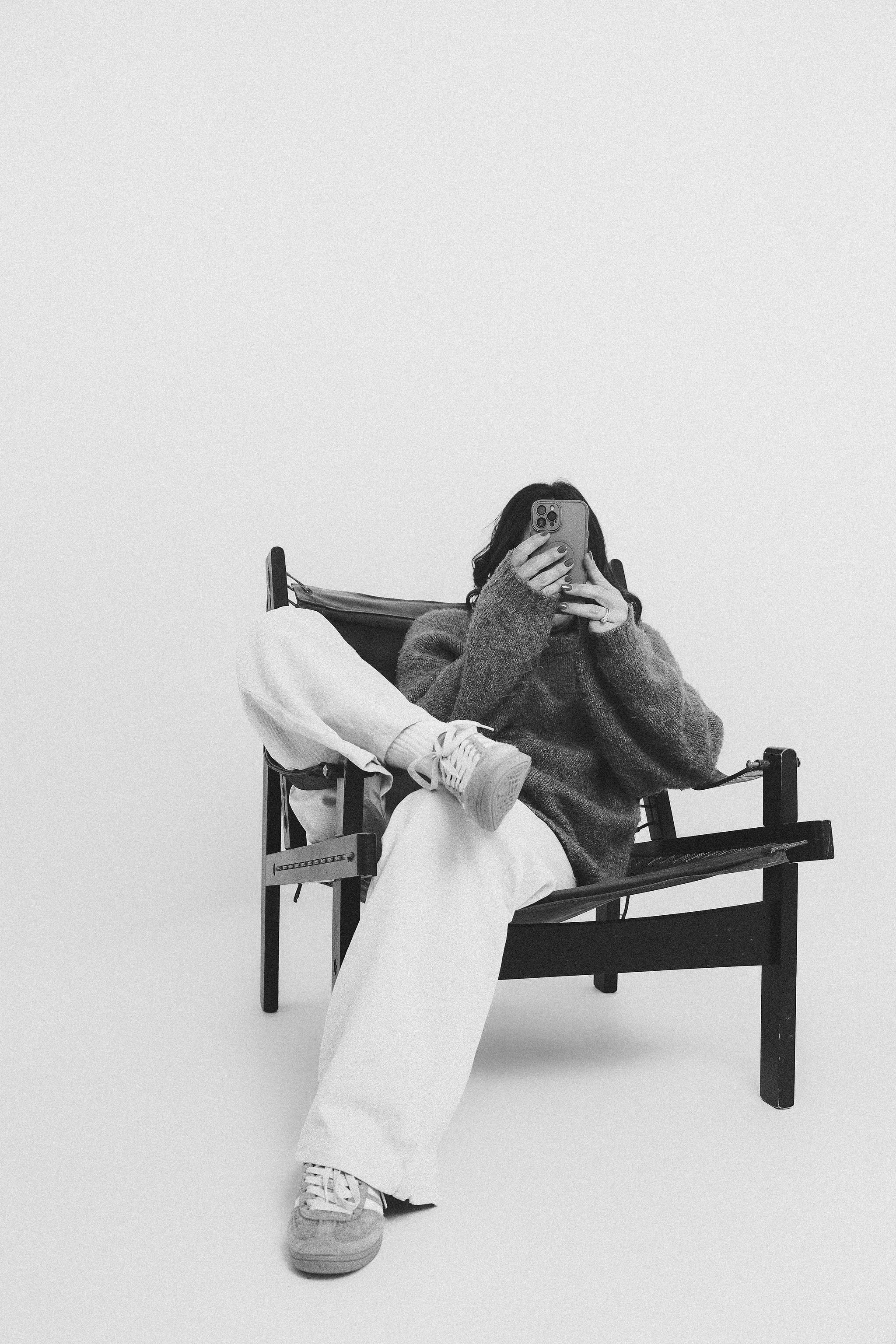 A woman sitting on a bench, taking a mirror selfie with her phone. She is wearing a thick sweater, loose white pants, and sneakers, with her hair partially visible.