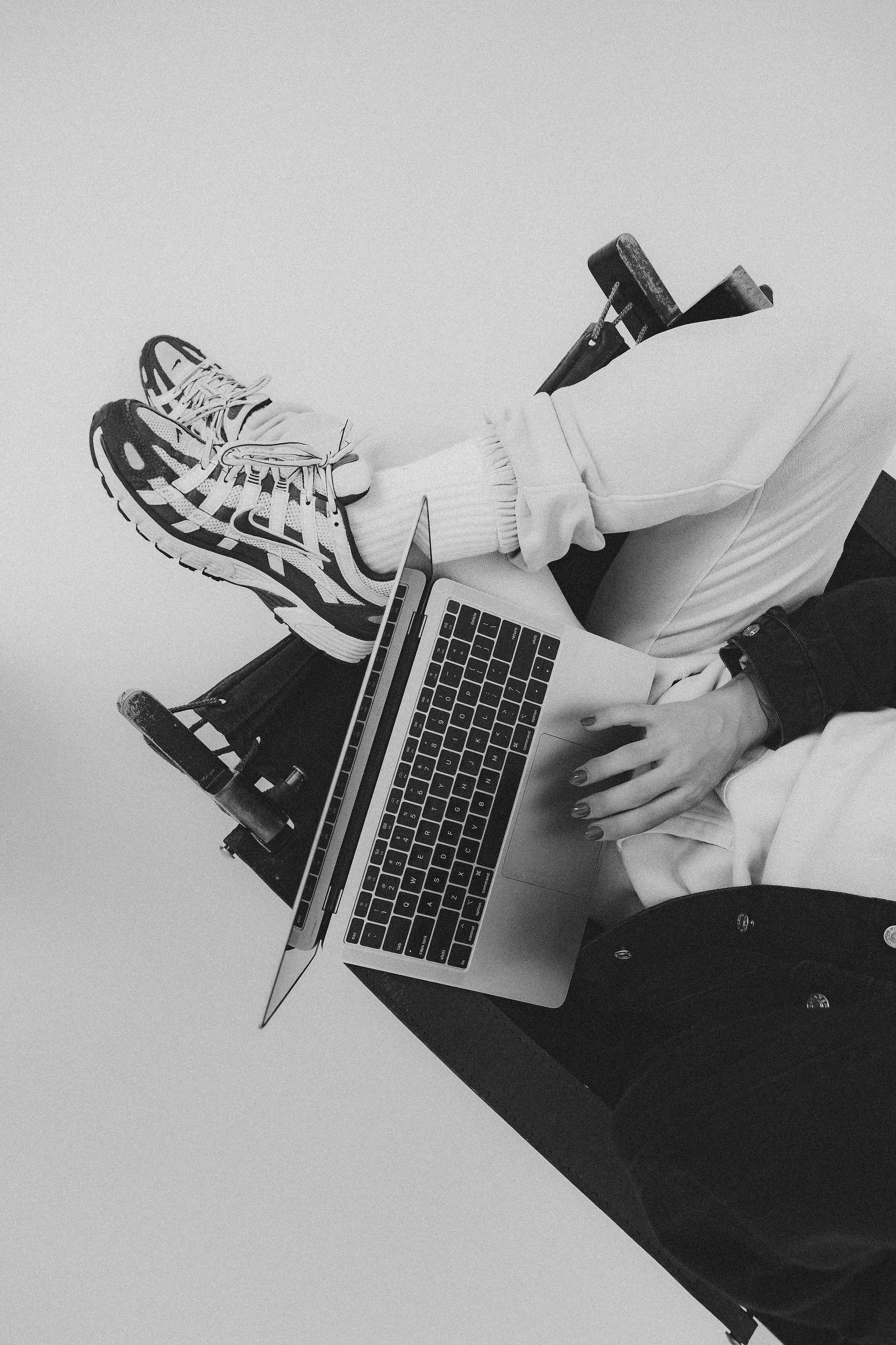 Person sitting with legs crossed, wearing sneakers and white pants, holding a laptop on lap. A hand resting near the laptop trackpad.