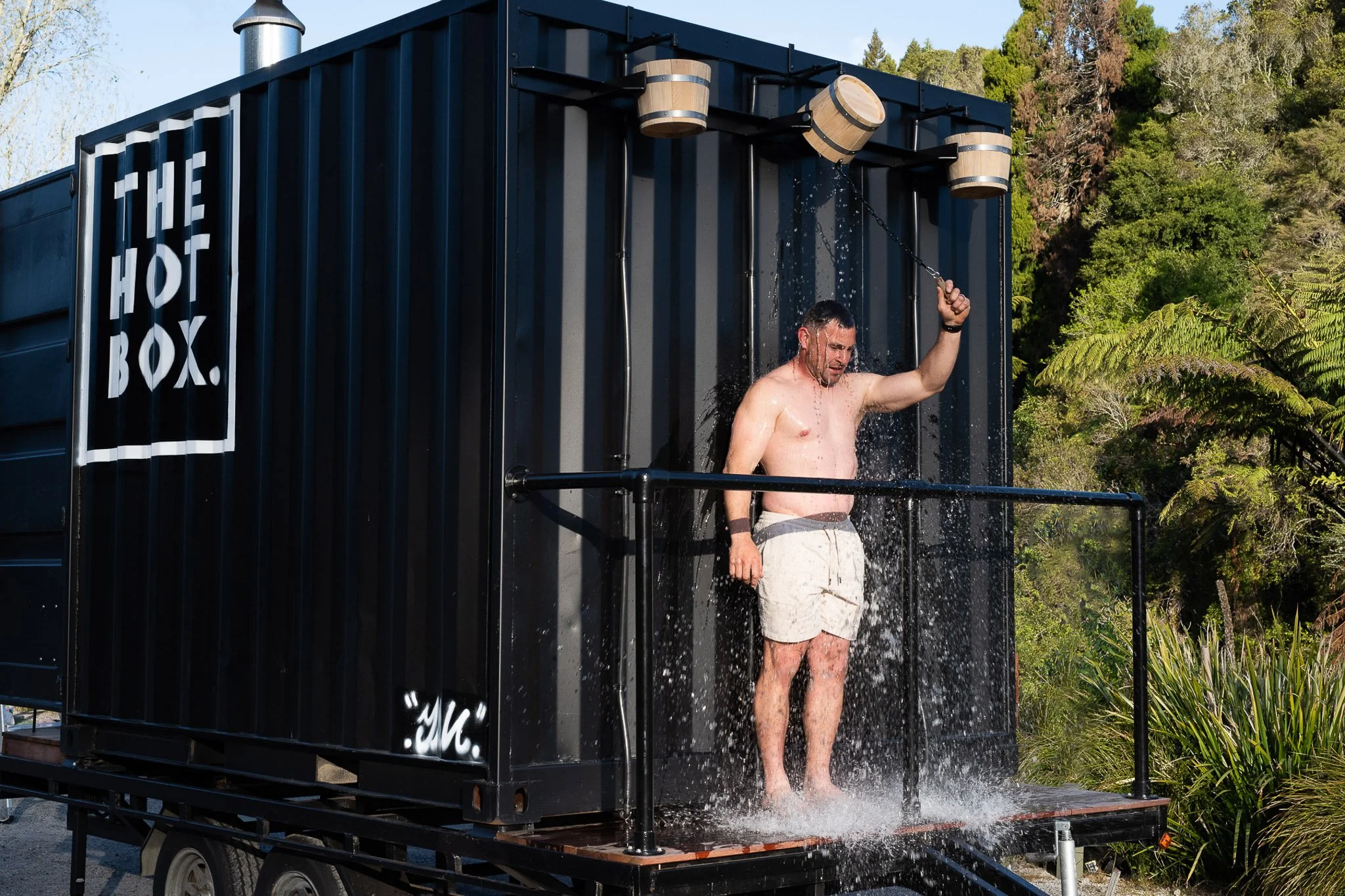 A man is taking a shower outside in a black, container-like structure labeled 'THE HOT BOX' with water running down. The structure is set outdoors with trees and greenery in the background.