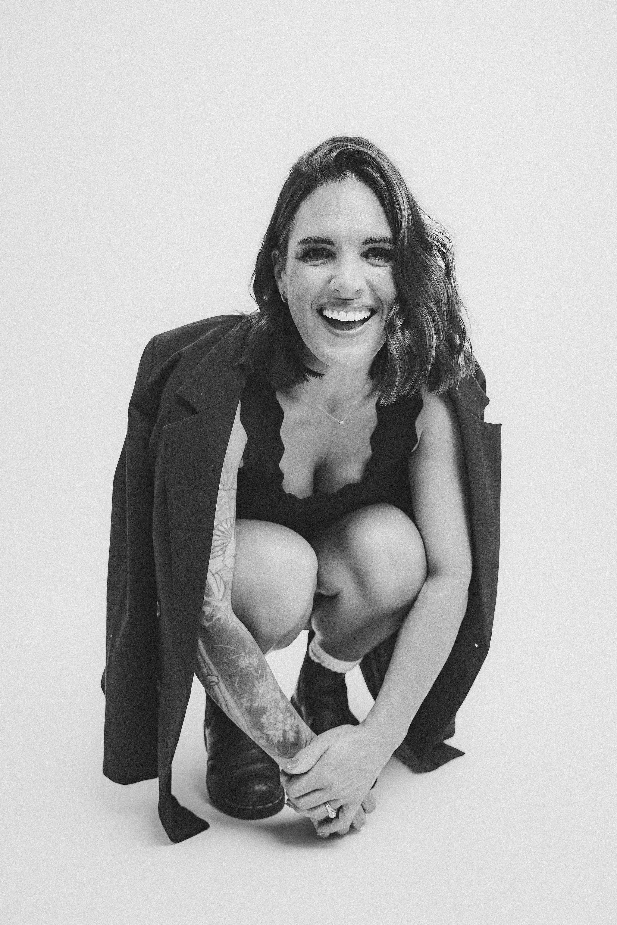 Black and white photo of a woman smiling, crouching with her knees up, wearing black shorts, boots, and a blazer draped over her shoulders, against a plain background.