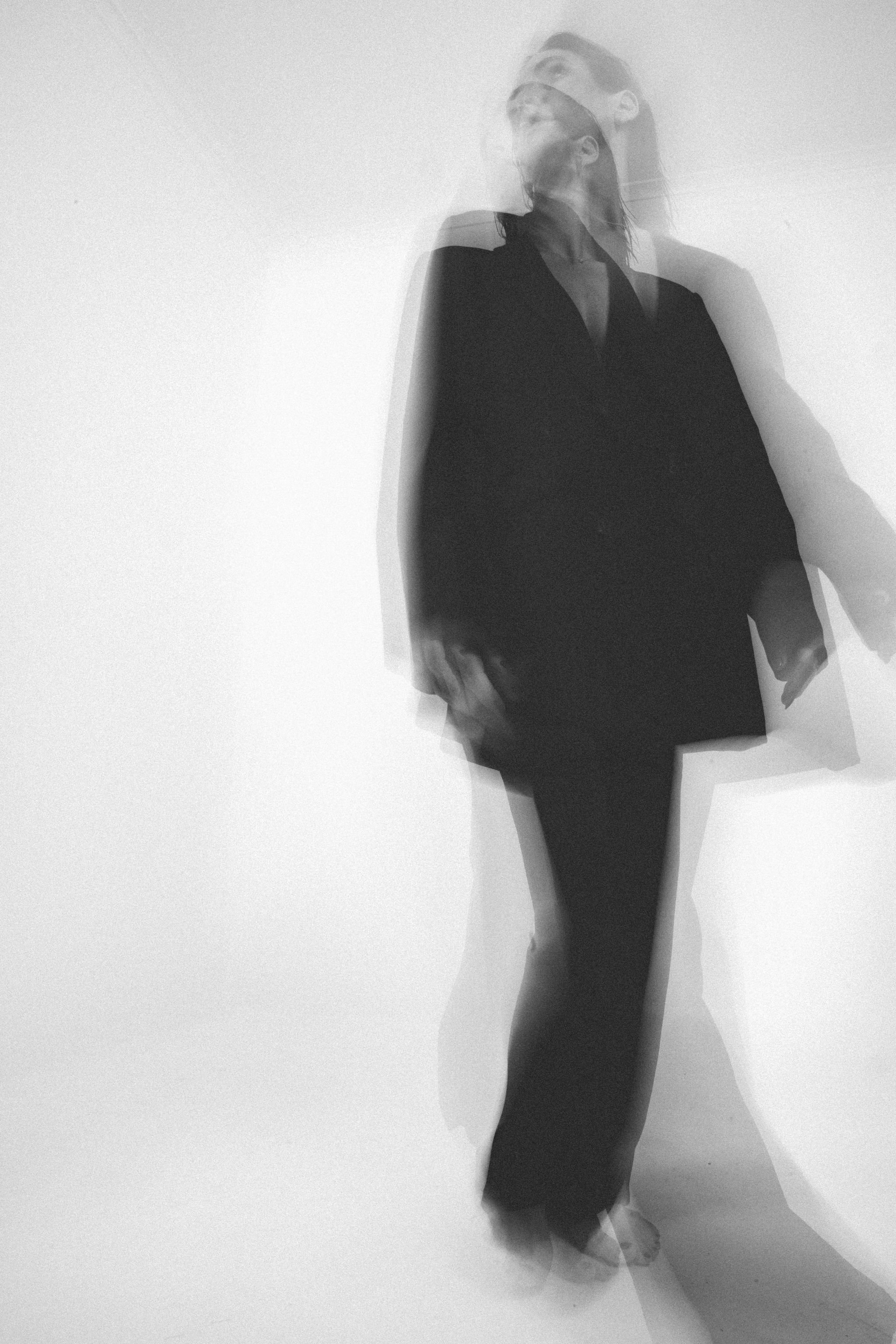 A black and white photo of a woman in a black outfit with a motion blur effect, creating multiple overlapping images of her smiling and looking to the side.