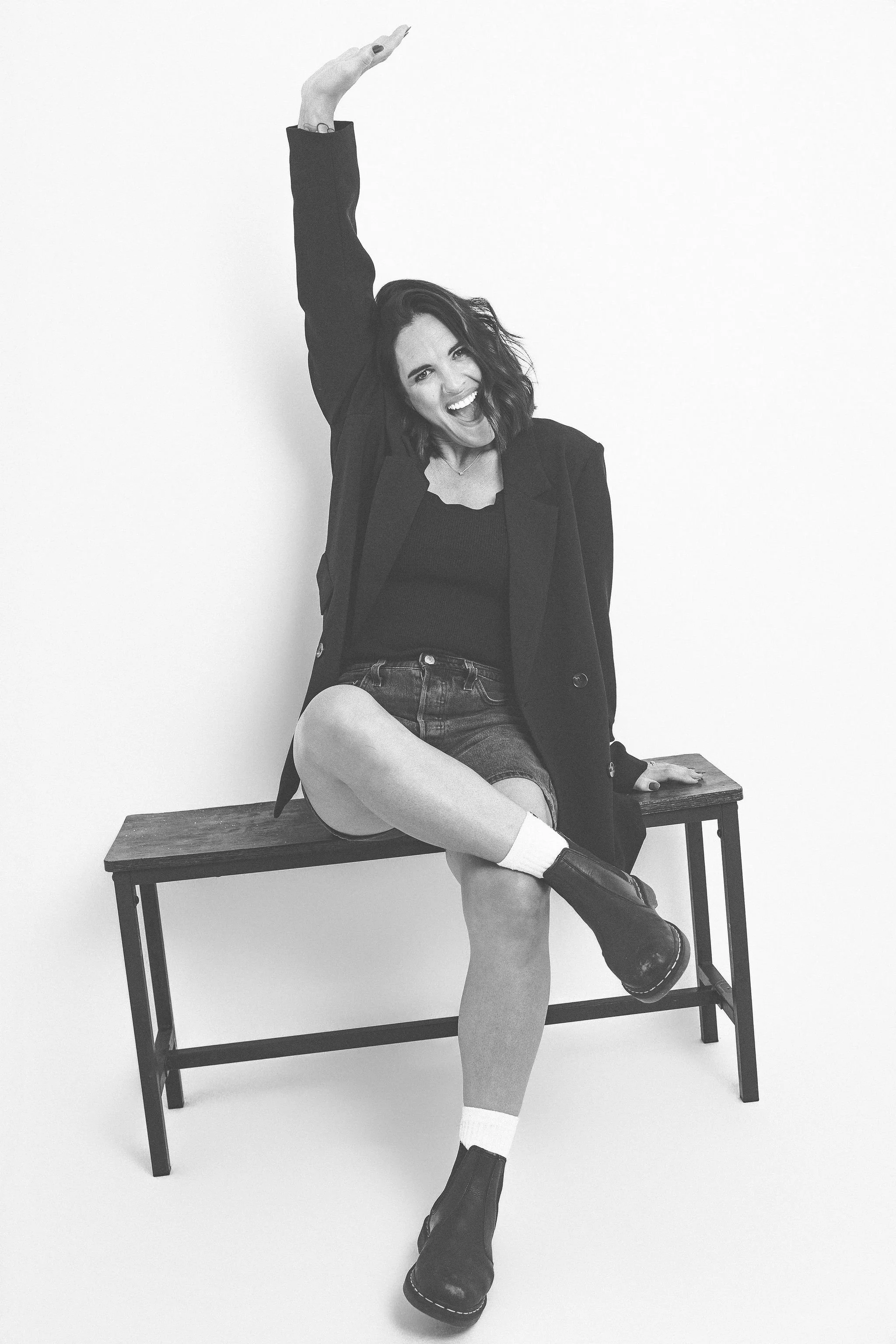 Black and white photo of a woman sitting on a wooden bench against a plain white background, smiling, with one arm raised and the other hand resting on the bench, wearing a blazer, t-shirt, shorts, socks, and boots.