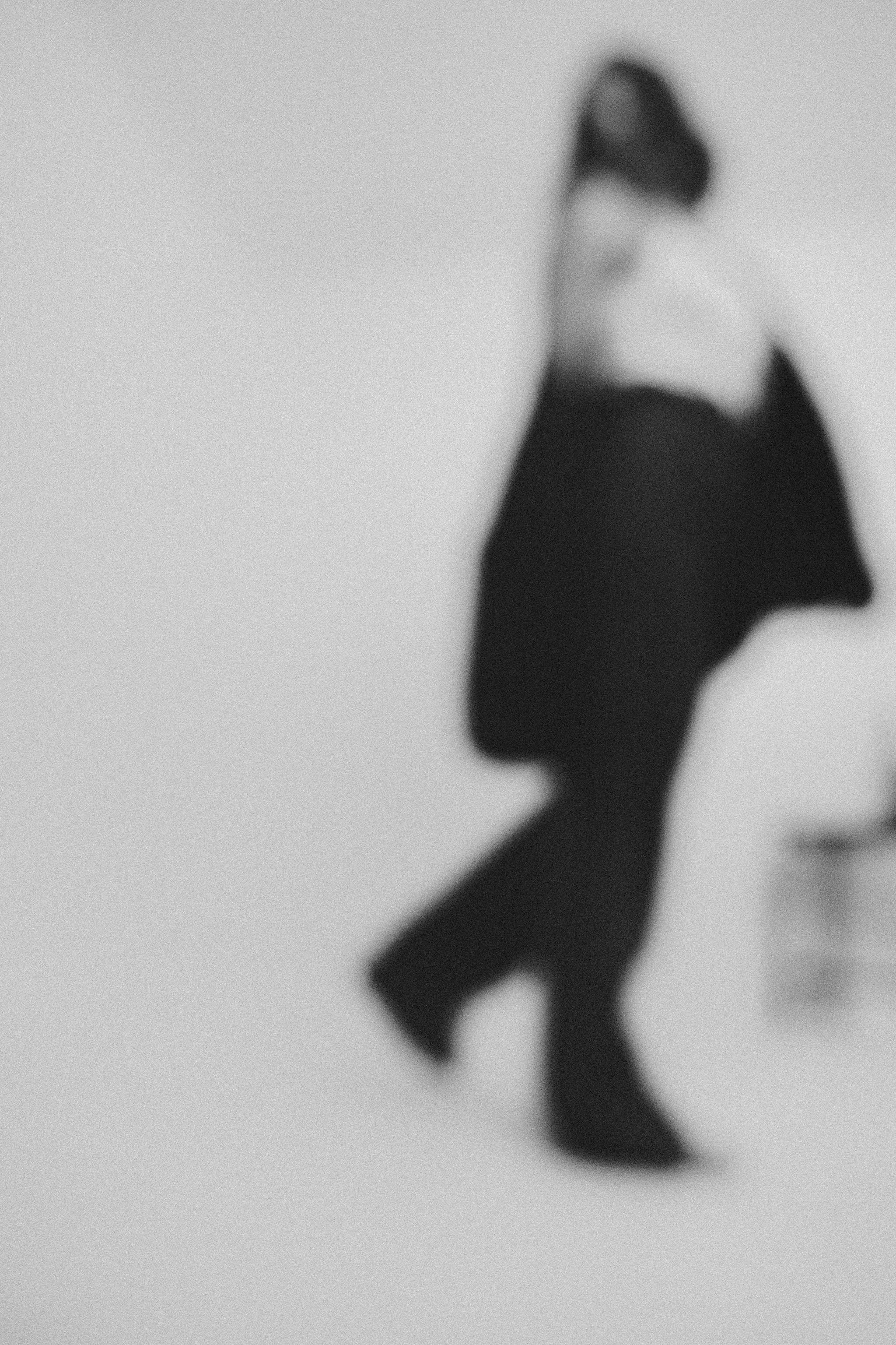 A blurred black and white photo of a person walking, wearing dark clothing, against a plain background.