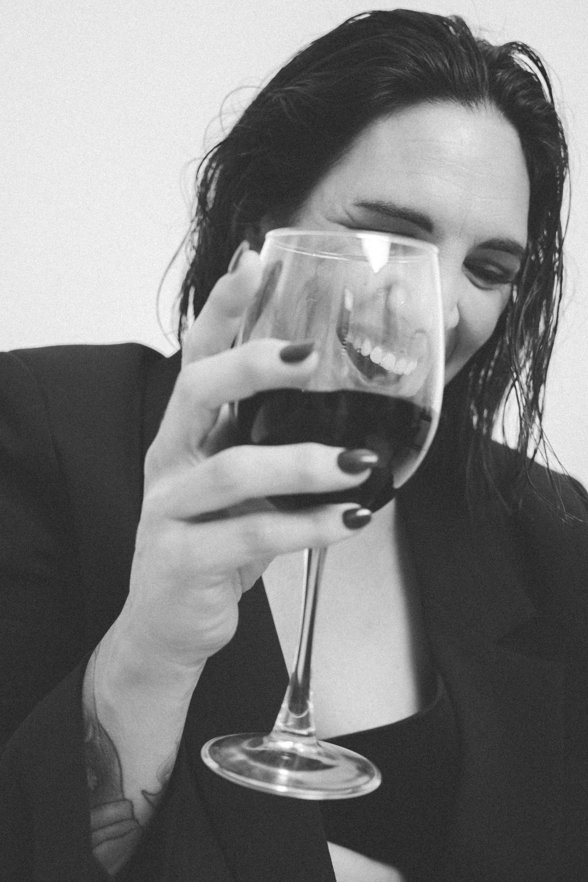 Black and white photo of a woman with dark hair smiling and holding a glass of red wine close to her face.