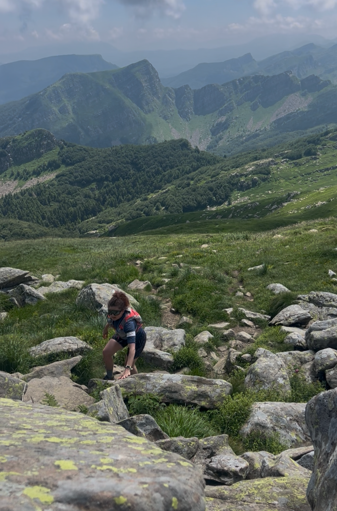 A young boy climbing over large rocks in a green mountainous landscape with distant mountains and a partly cloudy sky in the background.