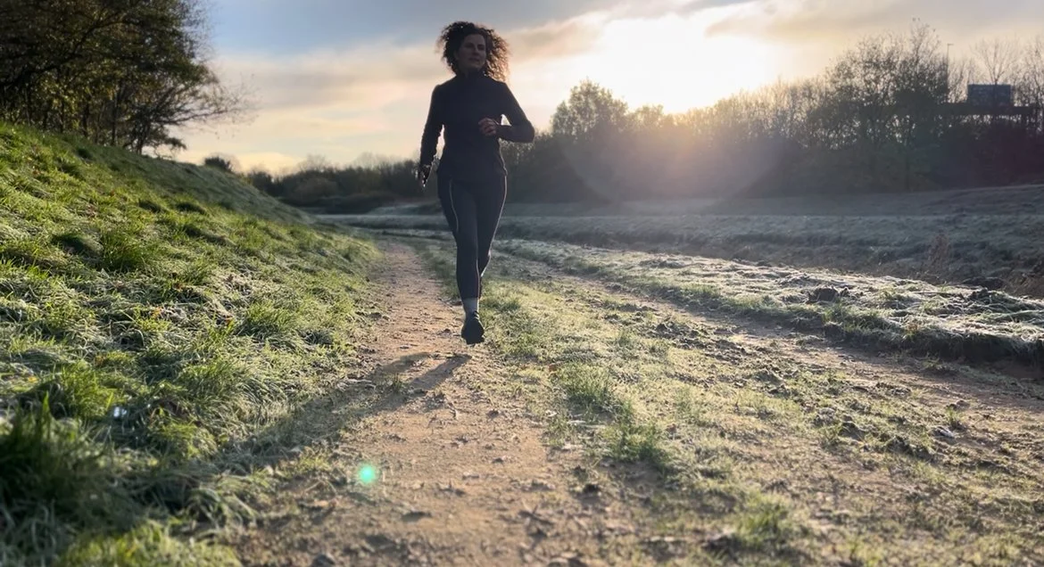 A woman jogging on a dirt path in a rural area during sunset, with trees in the background.
