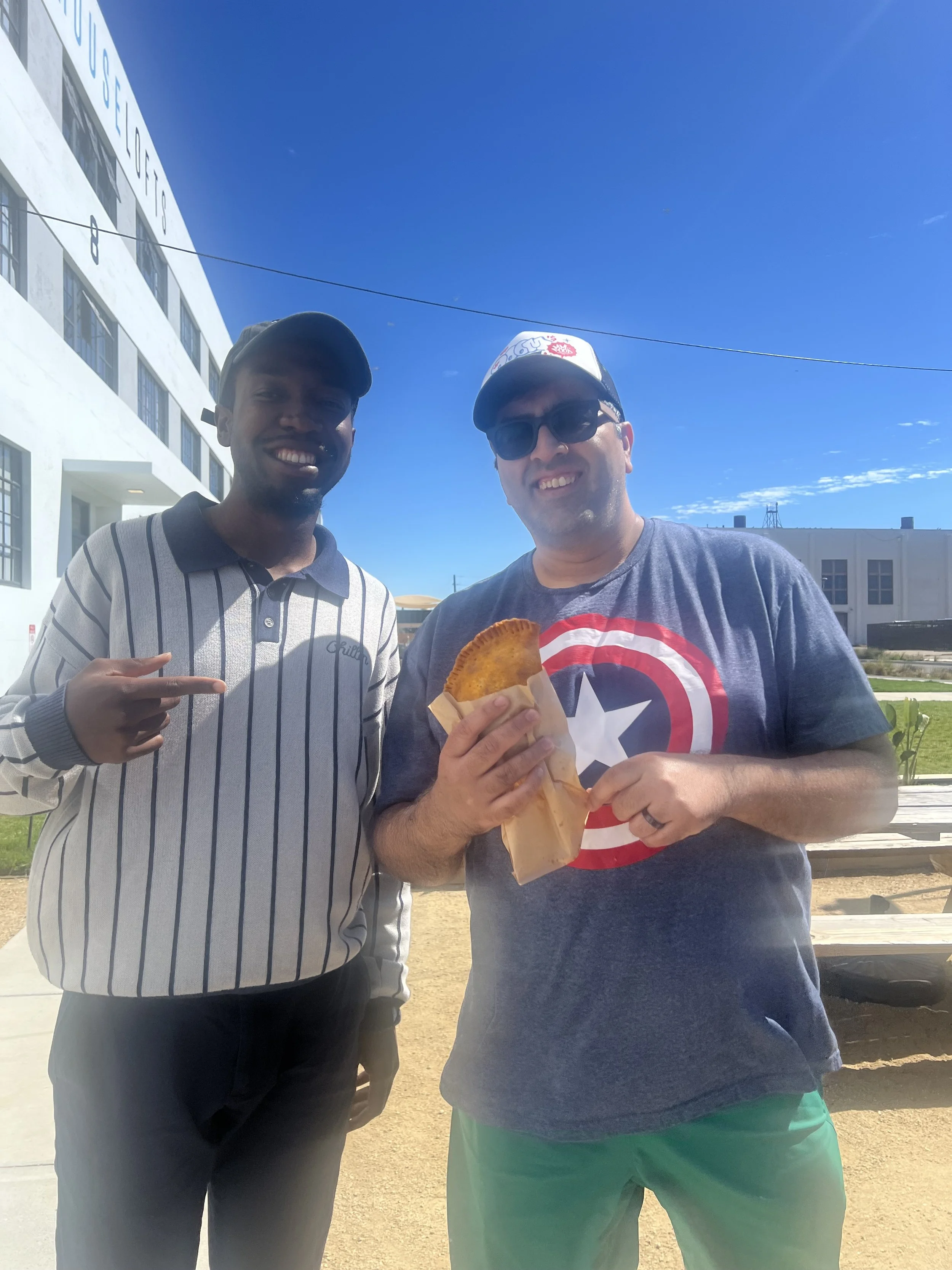 Two men standing outdoors under a clear blue sky. One man is pointing at the other, who is holding a pastry. They are smiling. Background includes a white building and some grassy area.