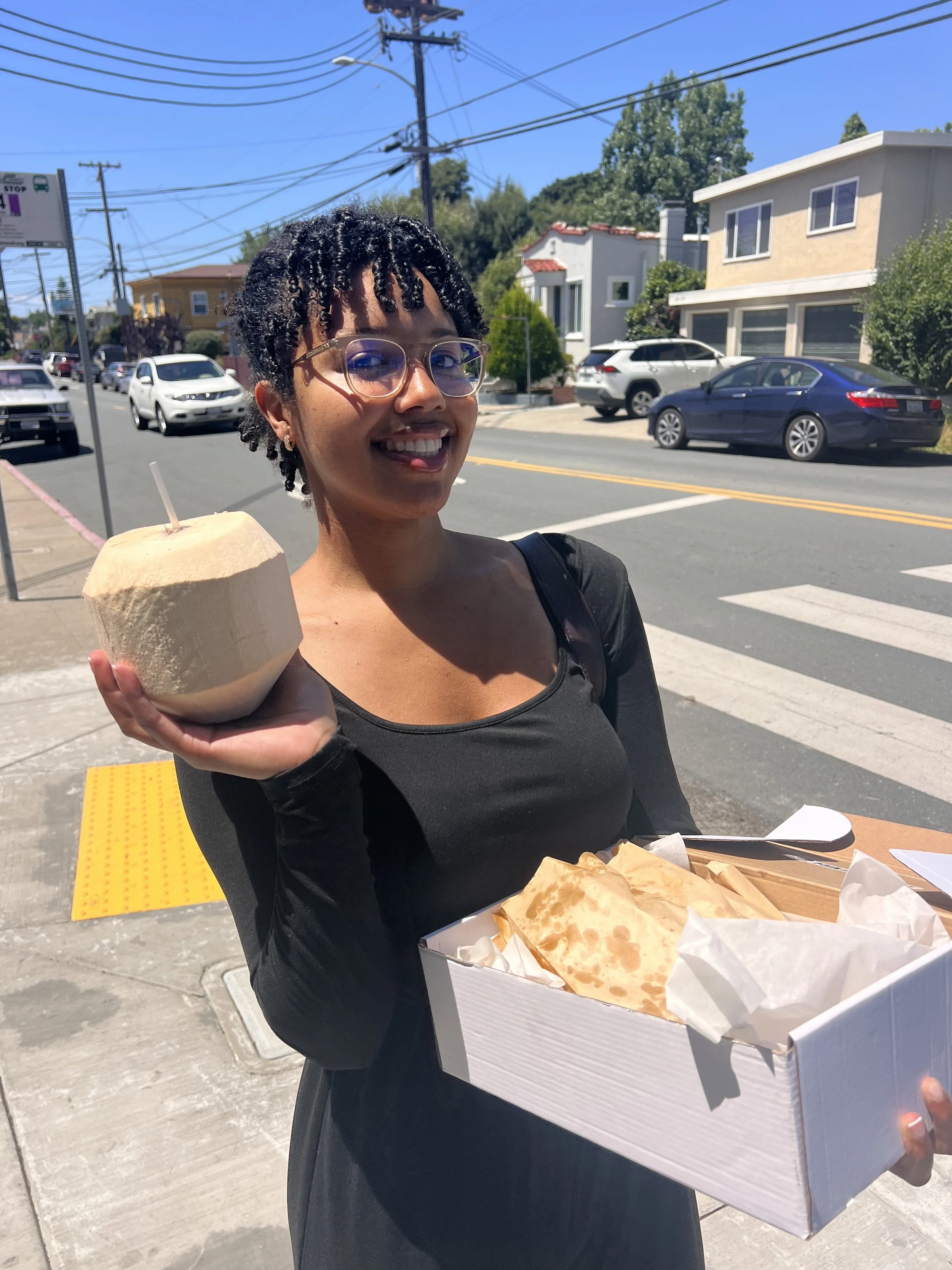 A smiling woman with glasses and short curly hair, holding a coconut in one hand and a box of food with paper wrappers in the other, standing on a city sidewalk on a sunny day.