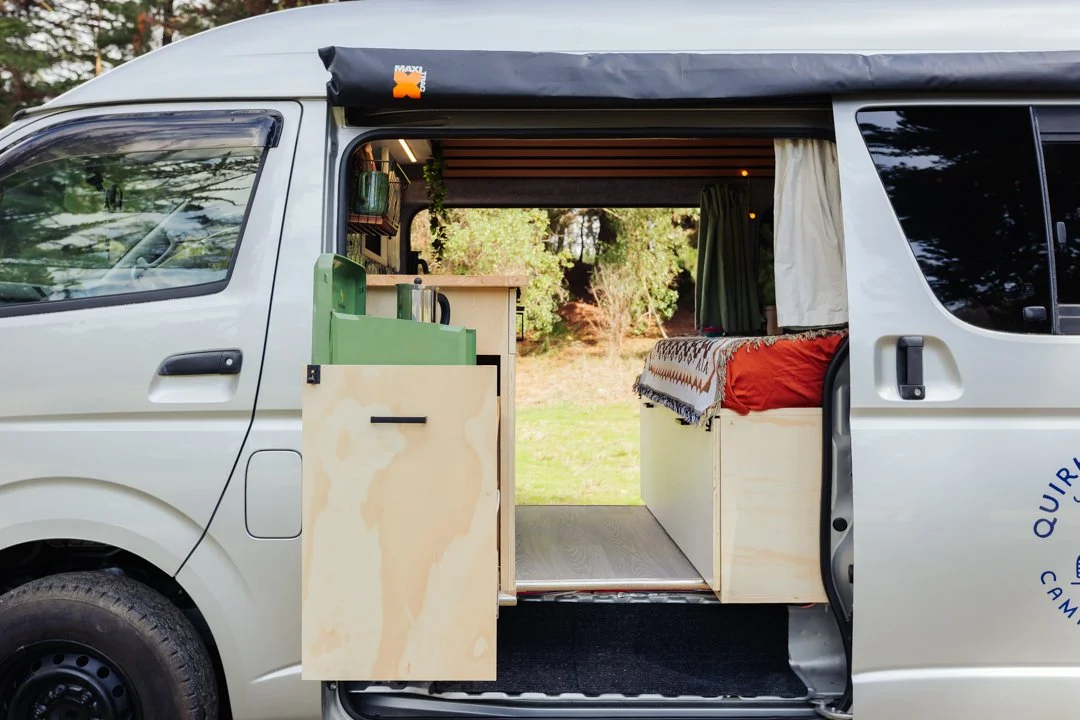 Interior view of a camper van with a bed, curtains, and a small kitchenette visible through an open side door.