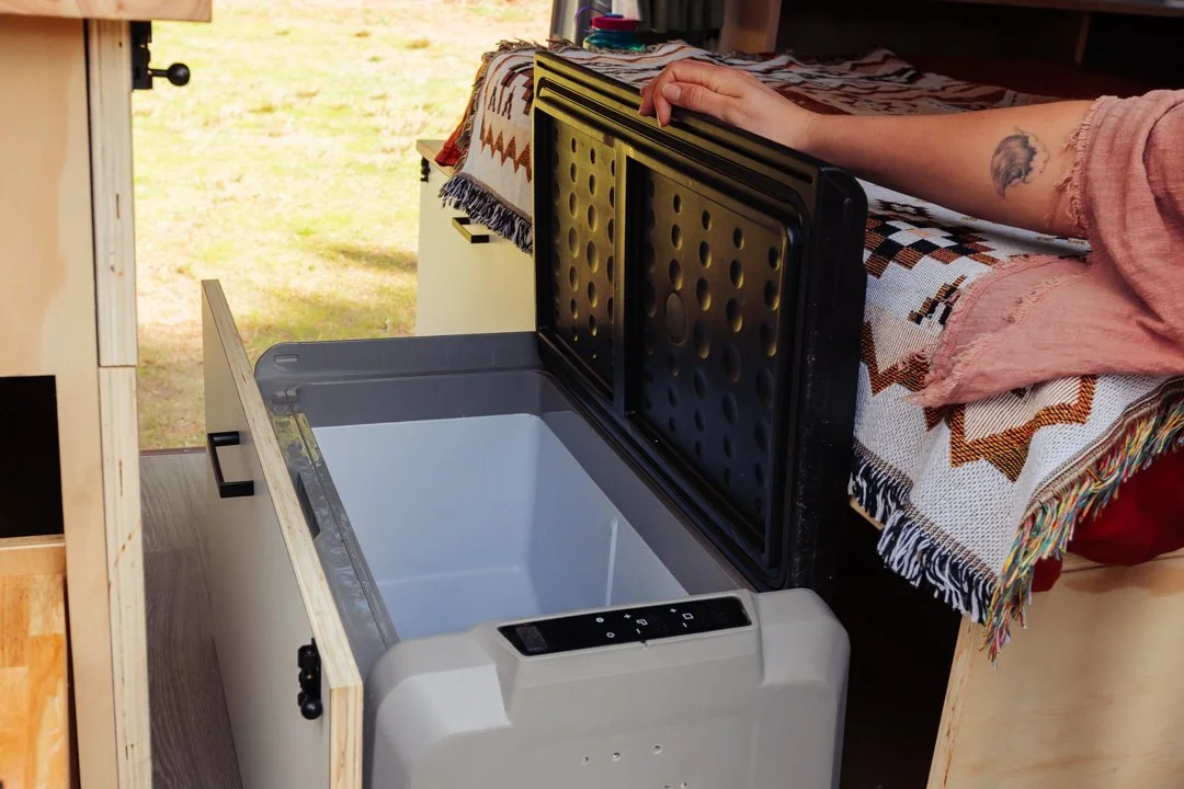 A person opening the lid of a white portable cooler in a cozy campervan conversion.