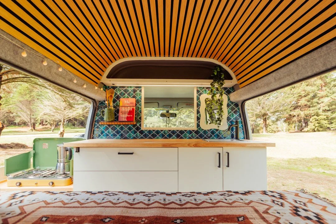 Interior of a camper van with a kitchen area, featuring a window, shelves with books and decor, a countertop, and a stove, set in a lush outdoor park.