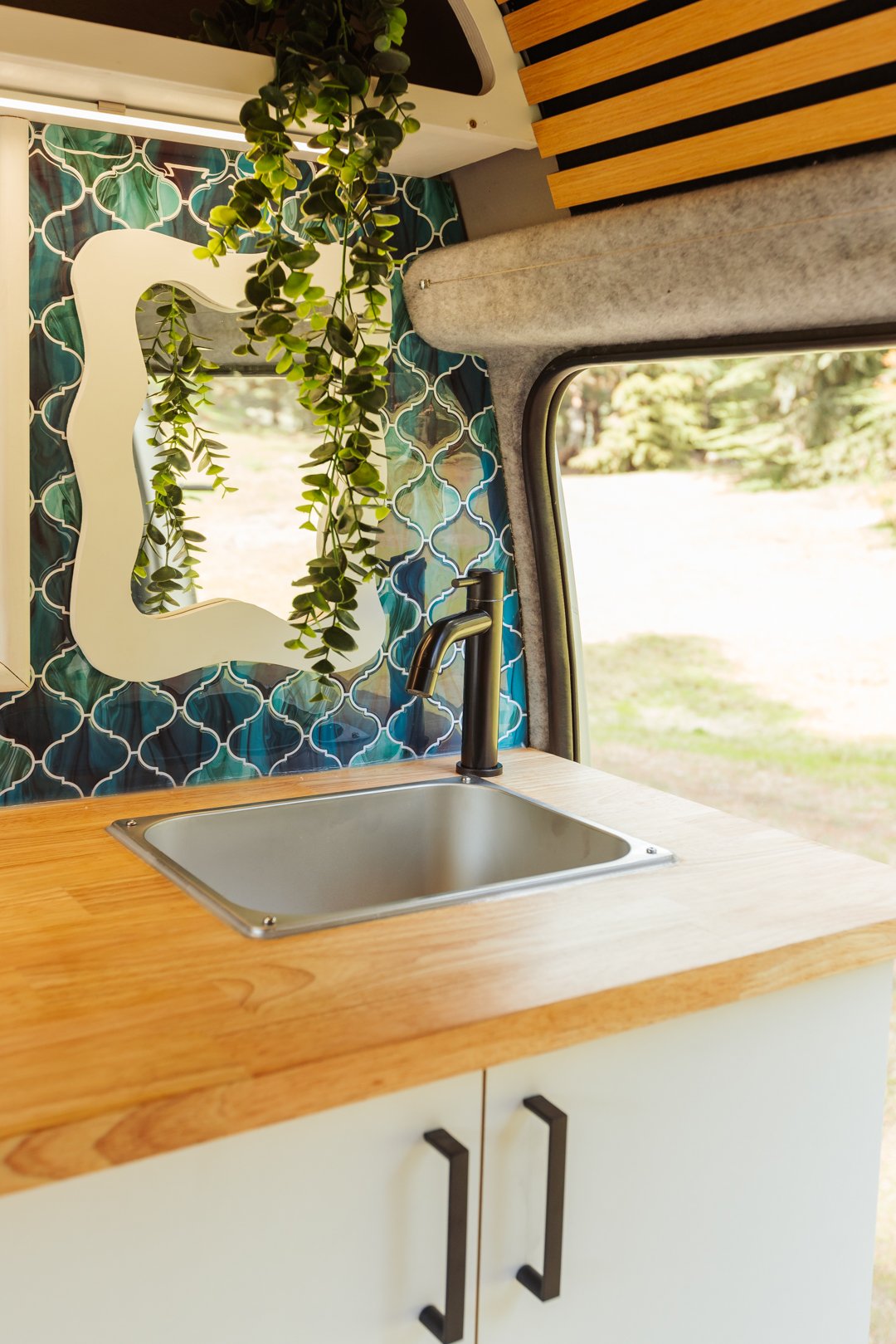 Interior of a van converted into a kitchen with a small sink, black faucet, wooden countertop, patterned blue tiles, mirror, hanging green plant, and window showing outdoor scenery.
