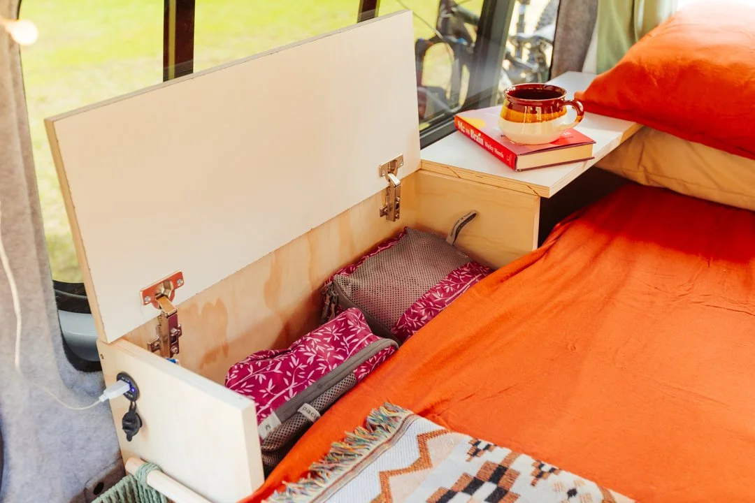A bed in a campervan, a small side table with a cup and a book, and a built-in storage compartment under the bed containing bags.