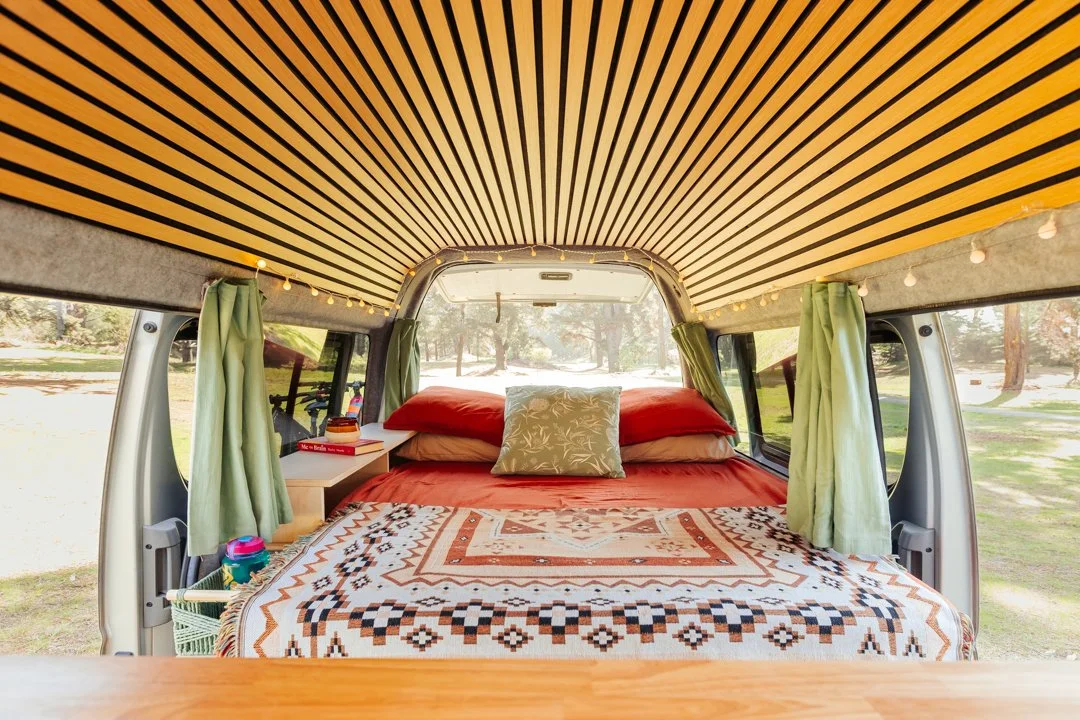 Inside a van converted into a camper with a bed, decorative quilt, pillows, and curtains, set in a park with trees visible through the windows.
