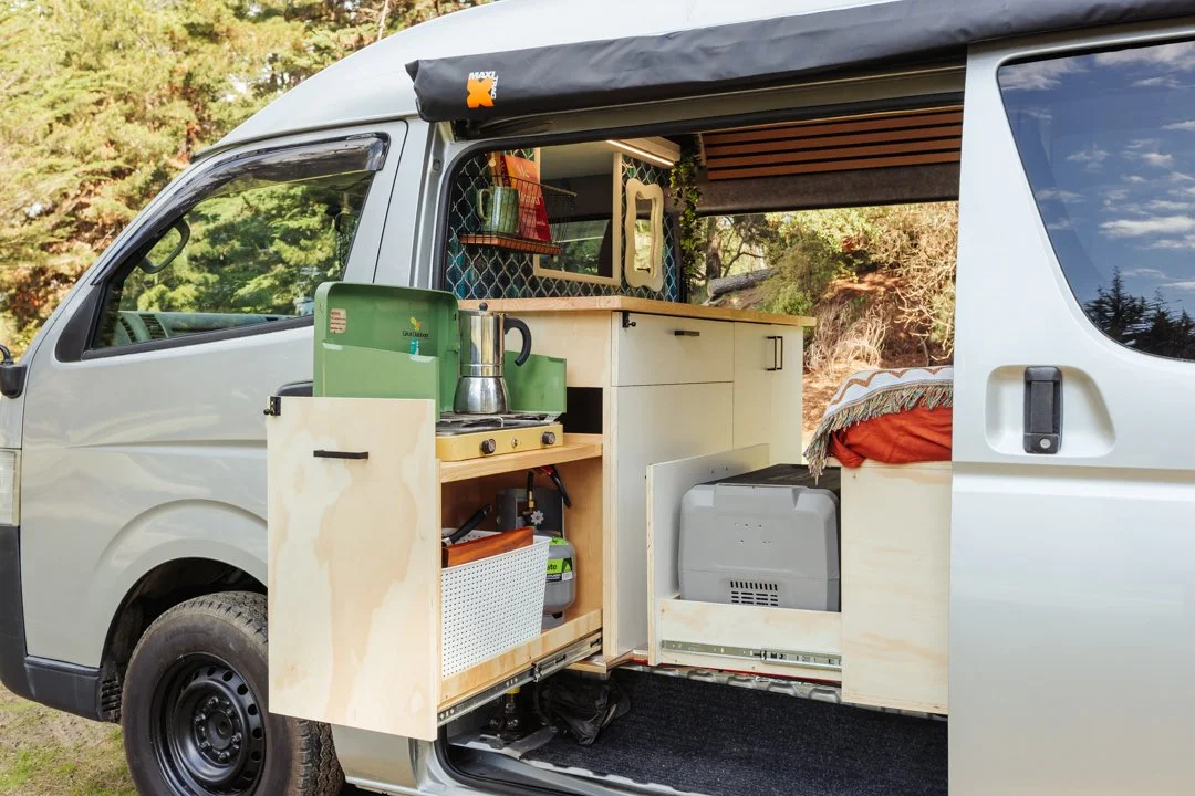 The inside of a camper van with a kitchenette, shelves, and bed, set in a natural outdoor environment.