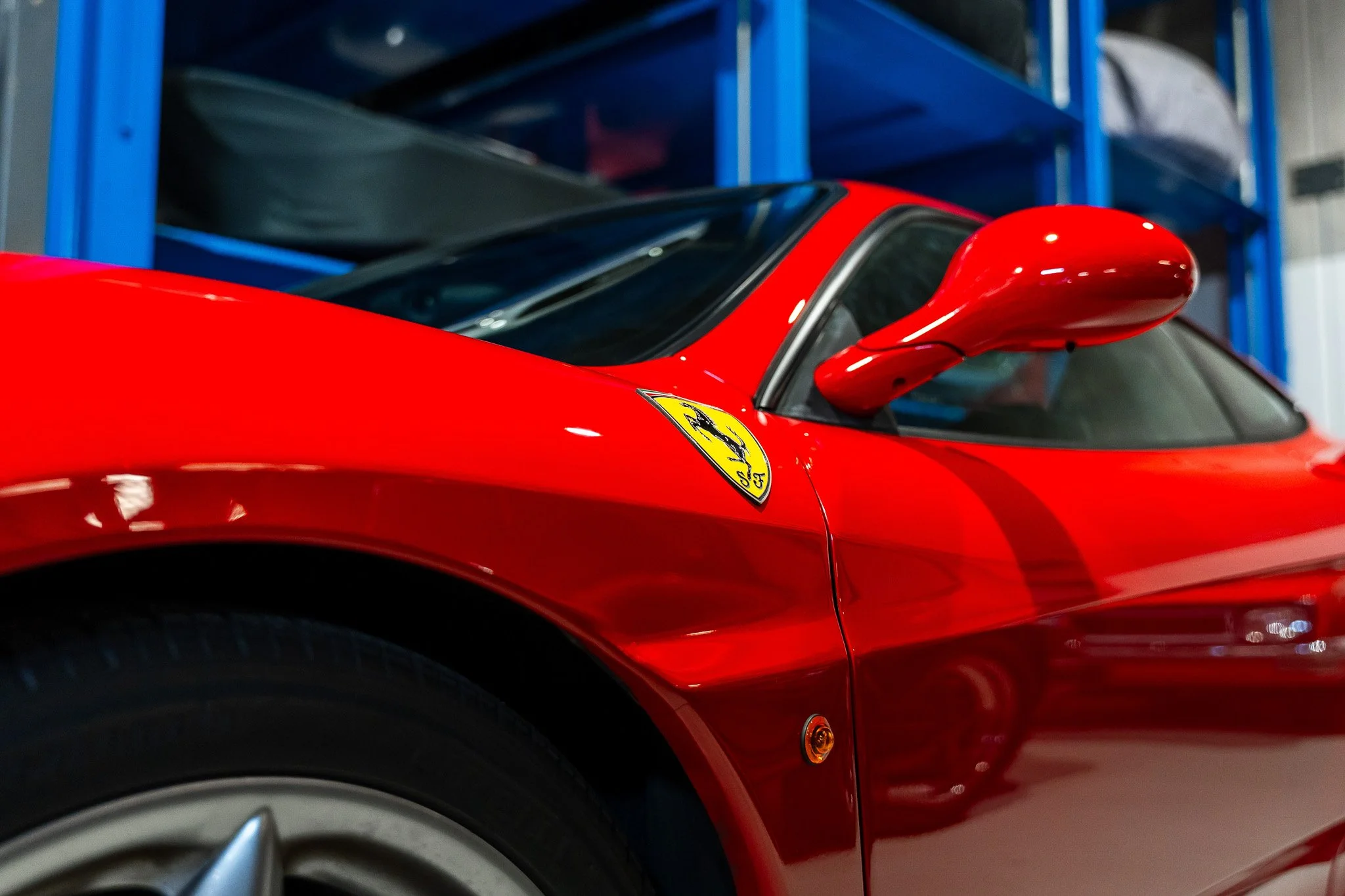 Close-up of a red sports car with a yellow Ferrari logo, parked indoors with blue storage shelves in the background.