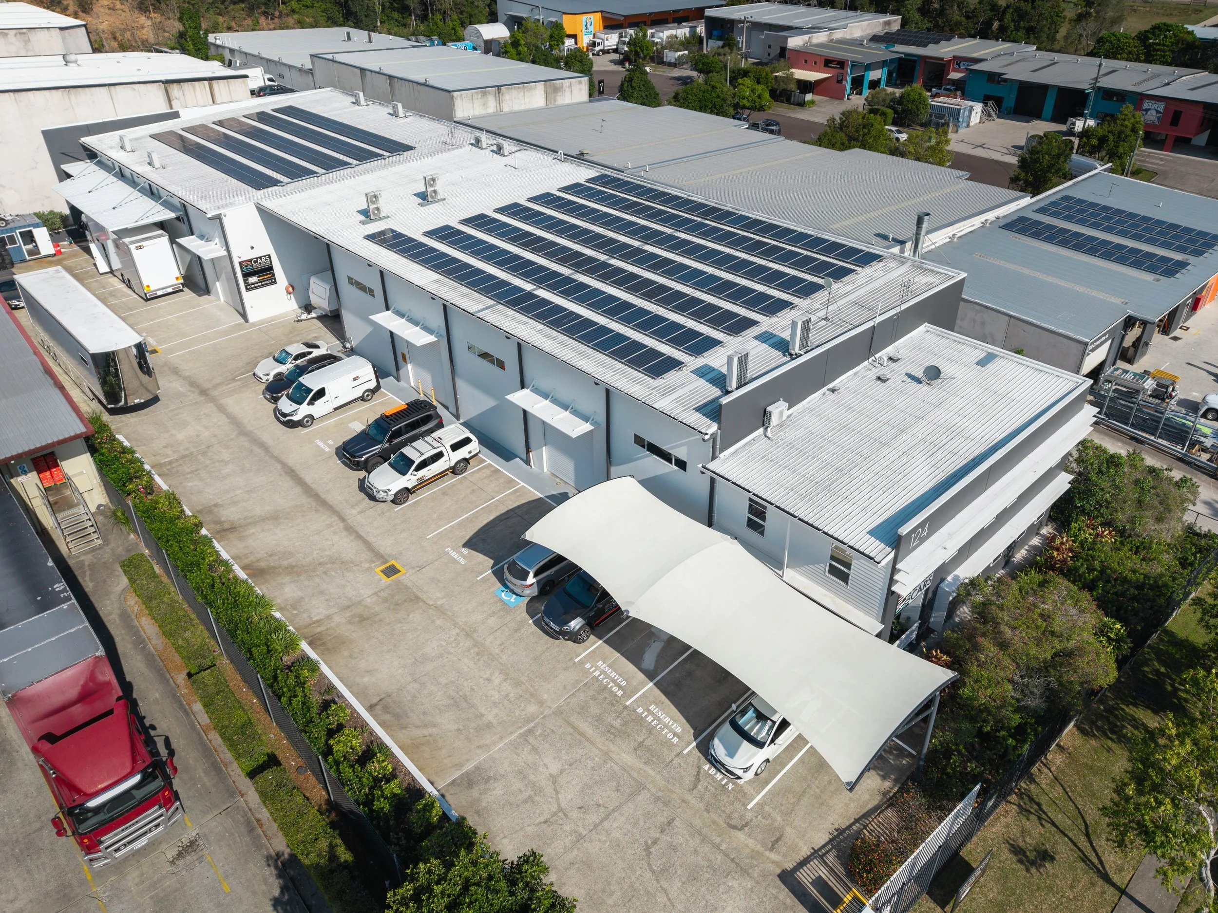 Aerial view of a commercial building with multiple solar panels on the roof, surrounded by parking lots with cars, trees, and adjacent buildings.