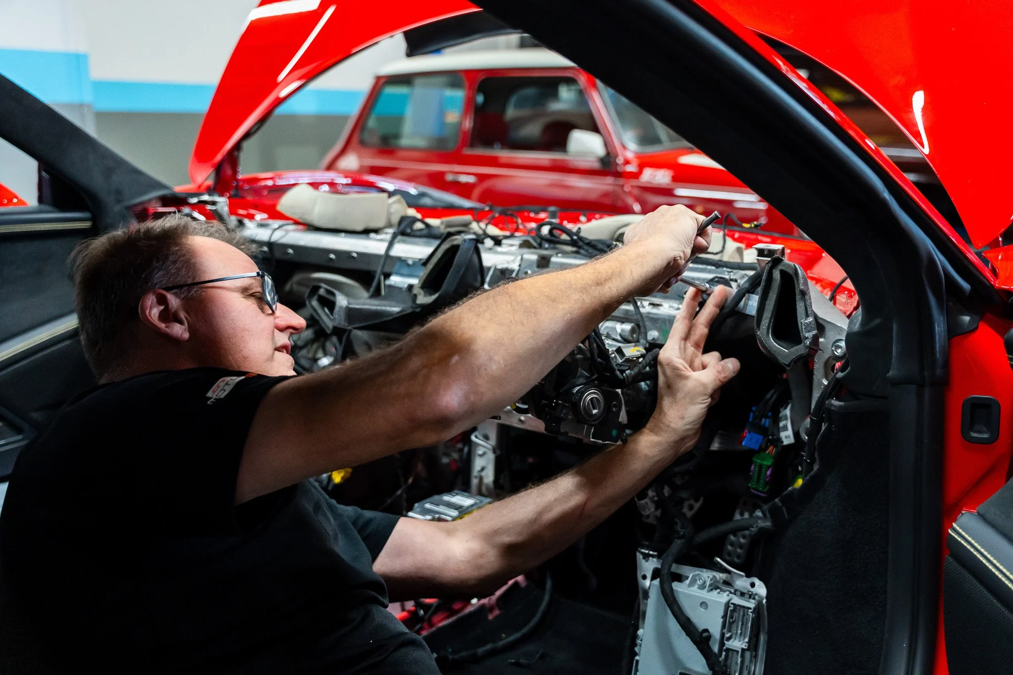 A man working on the internal engine of a red sports car in a workshop, with another red car visible in the background.