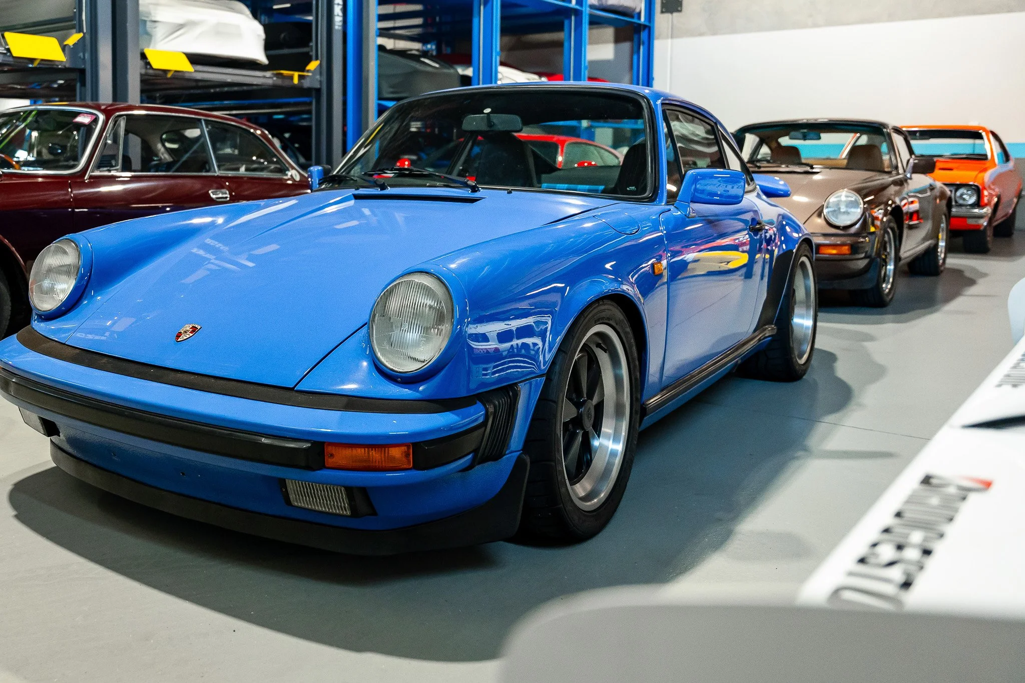 A blue vintage Porsche sports car parked indoors among other classic cars, with shelving units in the background holding various vehicle parts and items.