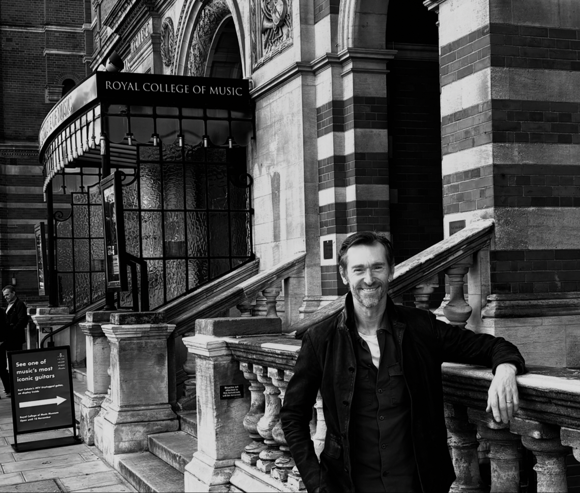 A man with a beard and dark hair, wearing a dark jacket, standing outside the Royal College of Music in London, smiling and leaning on a stone railing in front of a historic brick and stone building.