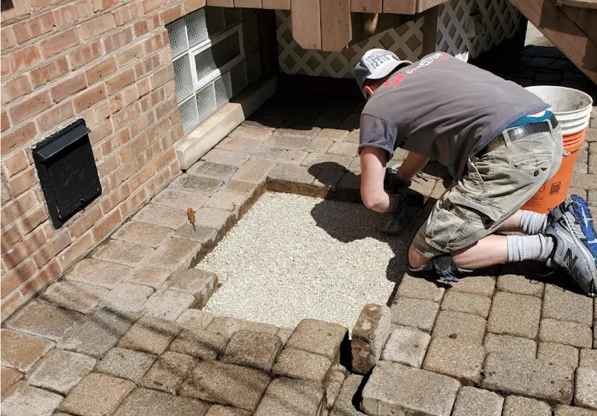 A man kneeling on a brick patio working on installing pavers with gravel base. He is wearing a gray t-shirt, shorts, and sneakers. There are buckets nearby and he is using his hands to place the pavers.