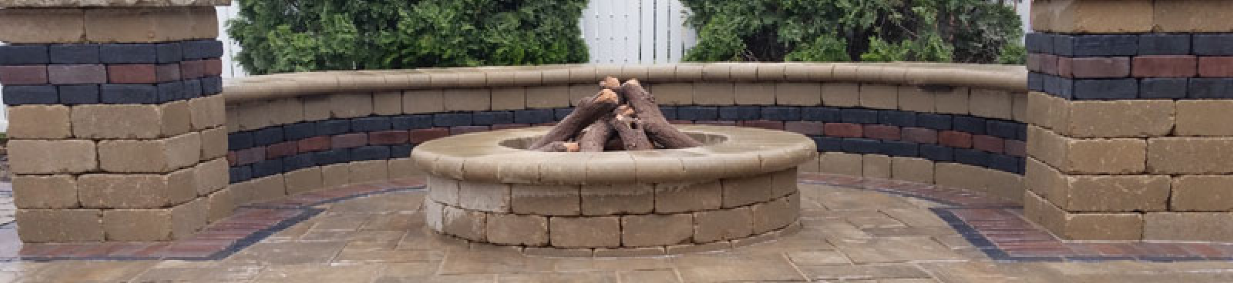 Brick fire pit with logs inside, surrounded by a brick wall with vertical colored bricks and pavers on the ground, with trees and a white fence in the background.