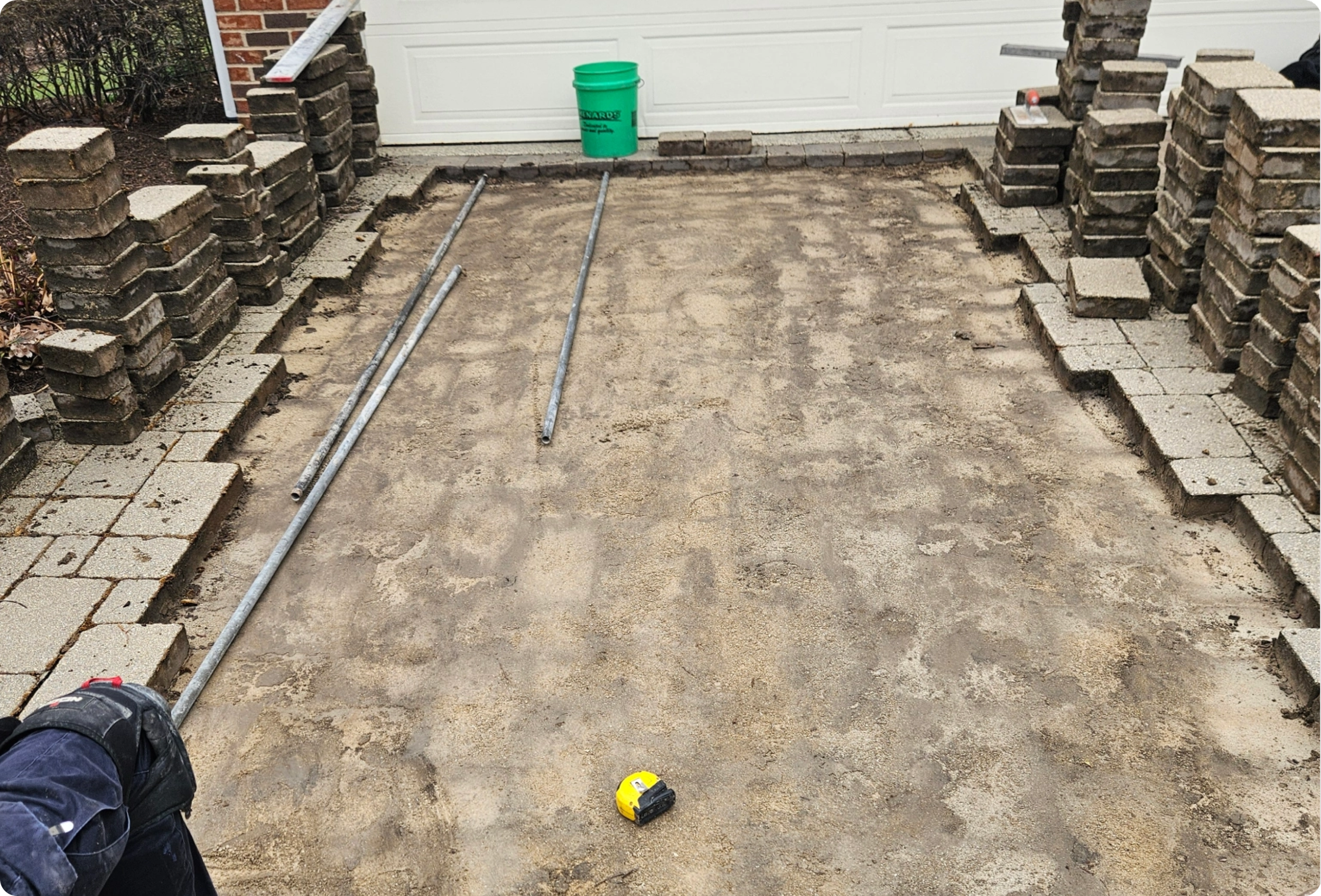 A driveway under construction with removed paving stones, metal pipes, a tape measure, and a green bucket near a garage door