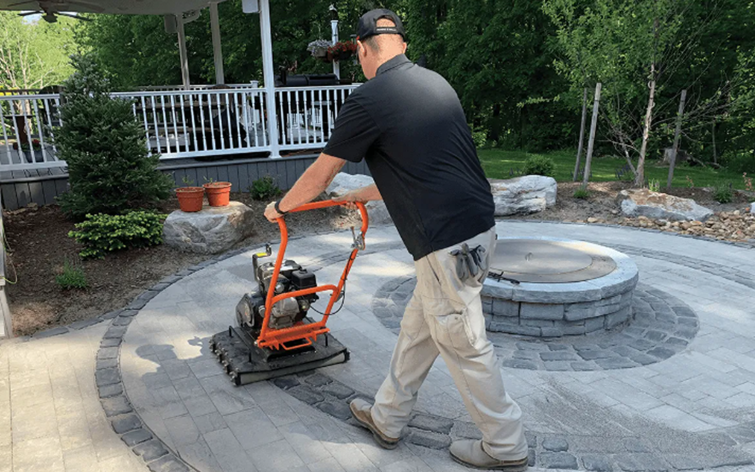 Man operating a plate compactor on a newly paved stone patio in a backyard, with trees, plants, and a raised deck in the background.
