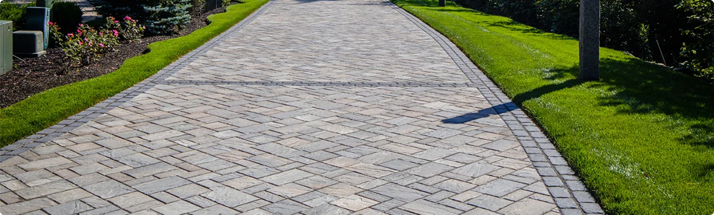 A paved walkway with interlocking stone bricks, bordered by green grass and a flower bed with pink flowers on the left side, and a manicured grassy area with trees on the right side.