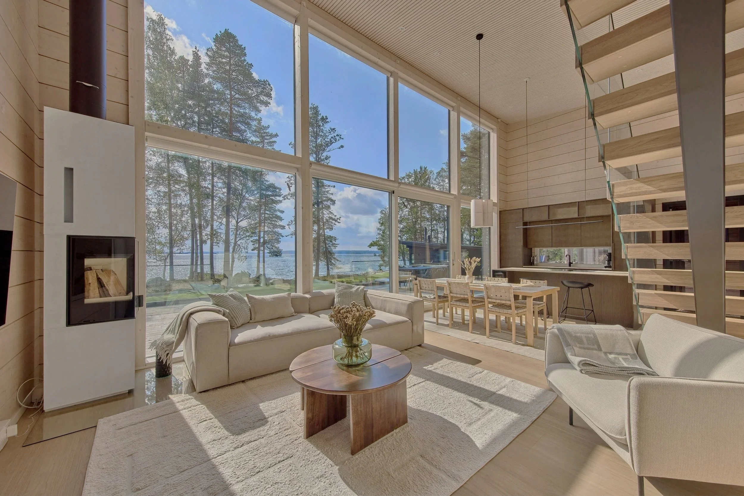 Avokeittiön ja olohuoneen avara sisätila suurella ikkunalla, näkymä järvelle ja puita, messinkinen työpöytä ja sohvakalusto.