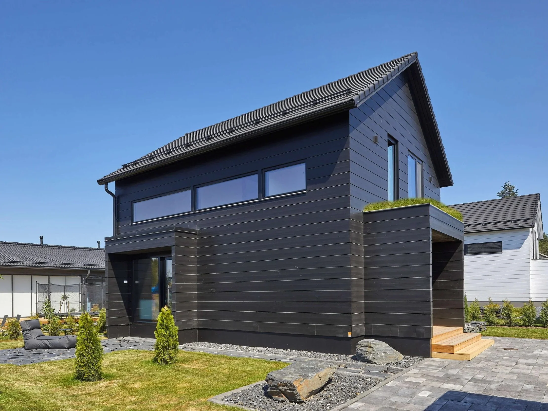 Modern mustavalkoinen talo pihalla, nurmikkoa ja kivipaasia edessä, taivas kirkas ja pilvetön.