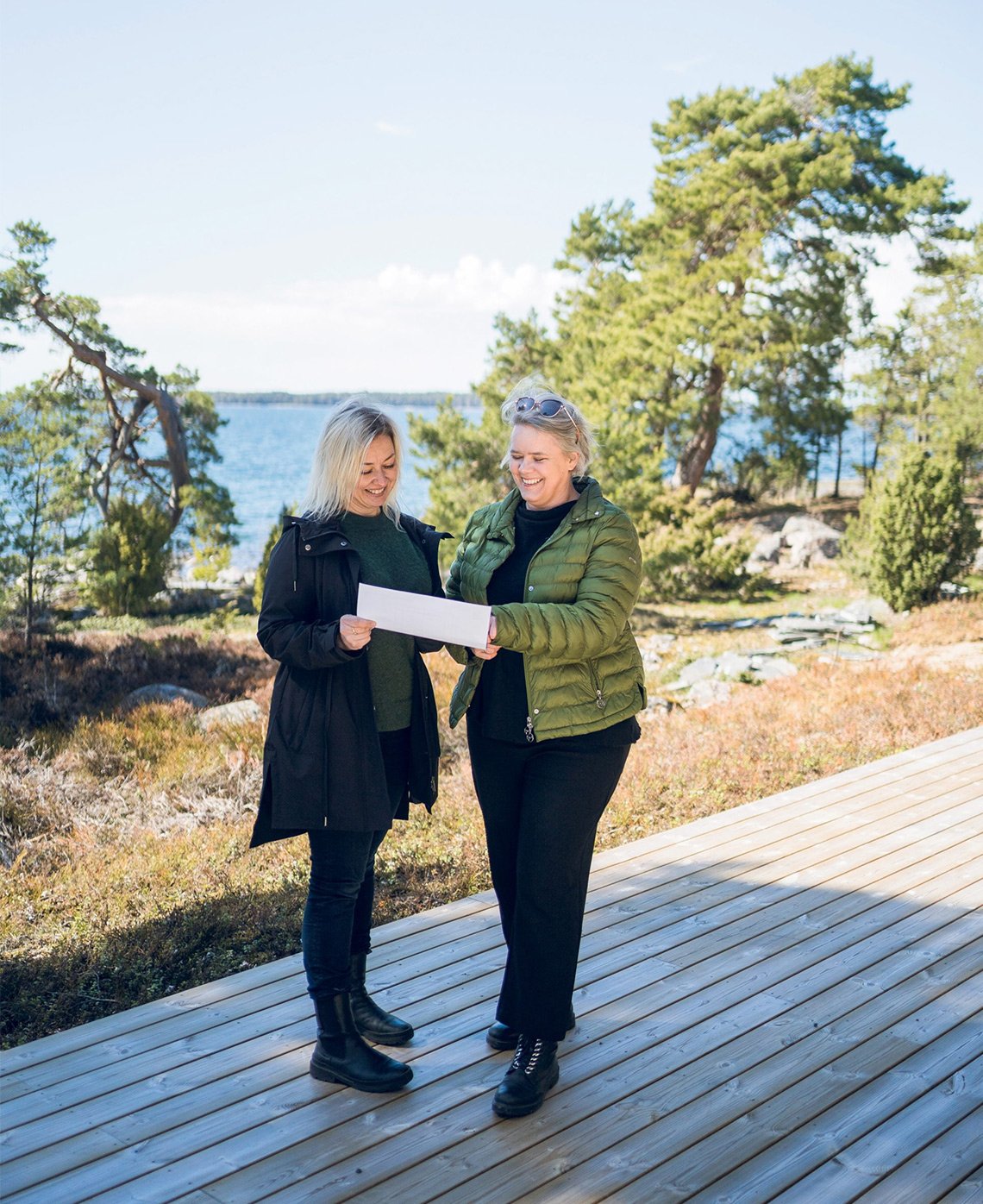 Kaksi naista seisoo laiturin päässä ja katselee pöydällä olevaa paperia meren ja puita sisältävän maiseman edessä.
