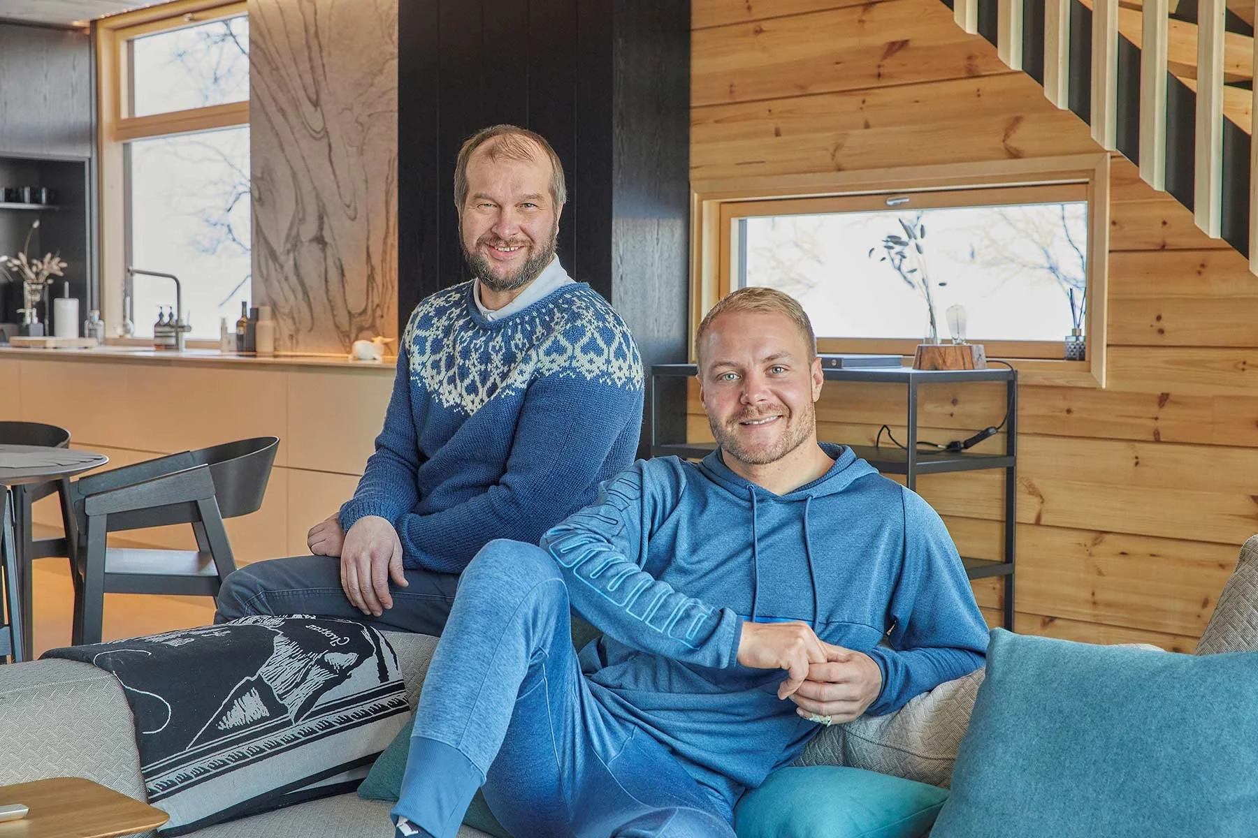 Kaksi miestä istuu ja seisoo modernissa asuintilassa, takana on puiset seinät ja suuri ikkuna, taustalla luonnon maisemaa.