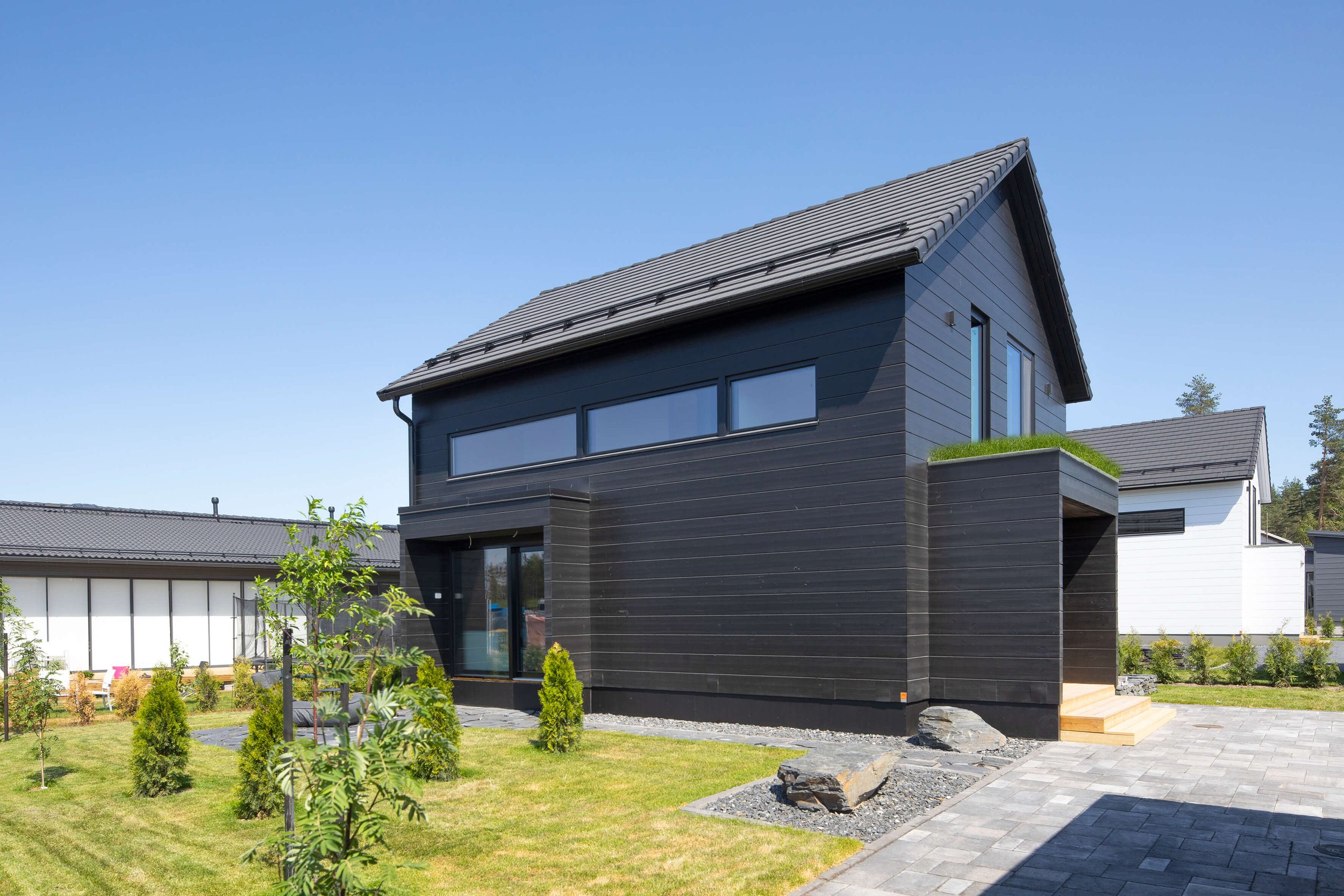 A view of the front yard of a modern black wooden building, with a lawn and plantings, and trees and a blue sky in the background.