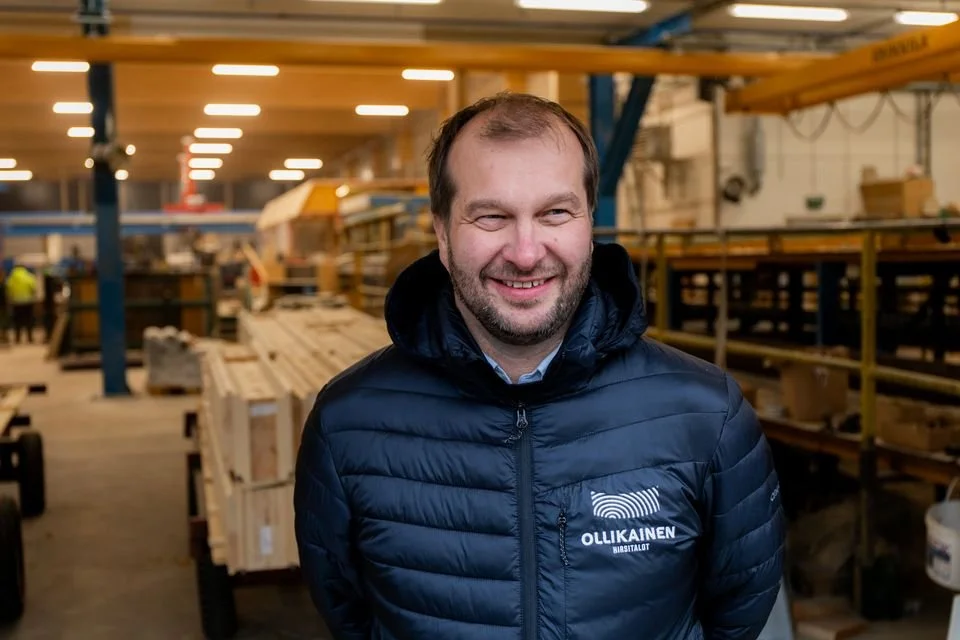 Ihmisen kasvot, hymyilevä mies sisällä tehdas- tai varastotilassa, takana on hyllyjä ja rakennustarvikkeita, päällä vaatteet, joissa on logo 'OLLKAISEN'.