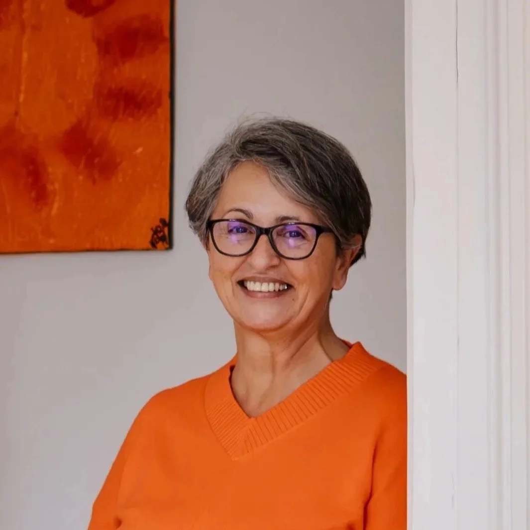 Eine lächelnde Frau mit kurzer grauer Frisur, Brille und orangem Pullover steht vor einer weißen Tür, im Hintergrund eine orange Wand mit Kunstwerk.