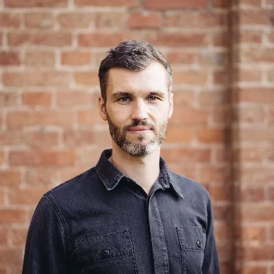 Porträt eines Mannes mit dunklem Hemd vor einer Ziegelsteinwand.